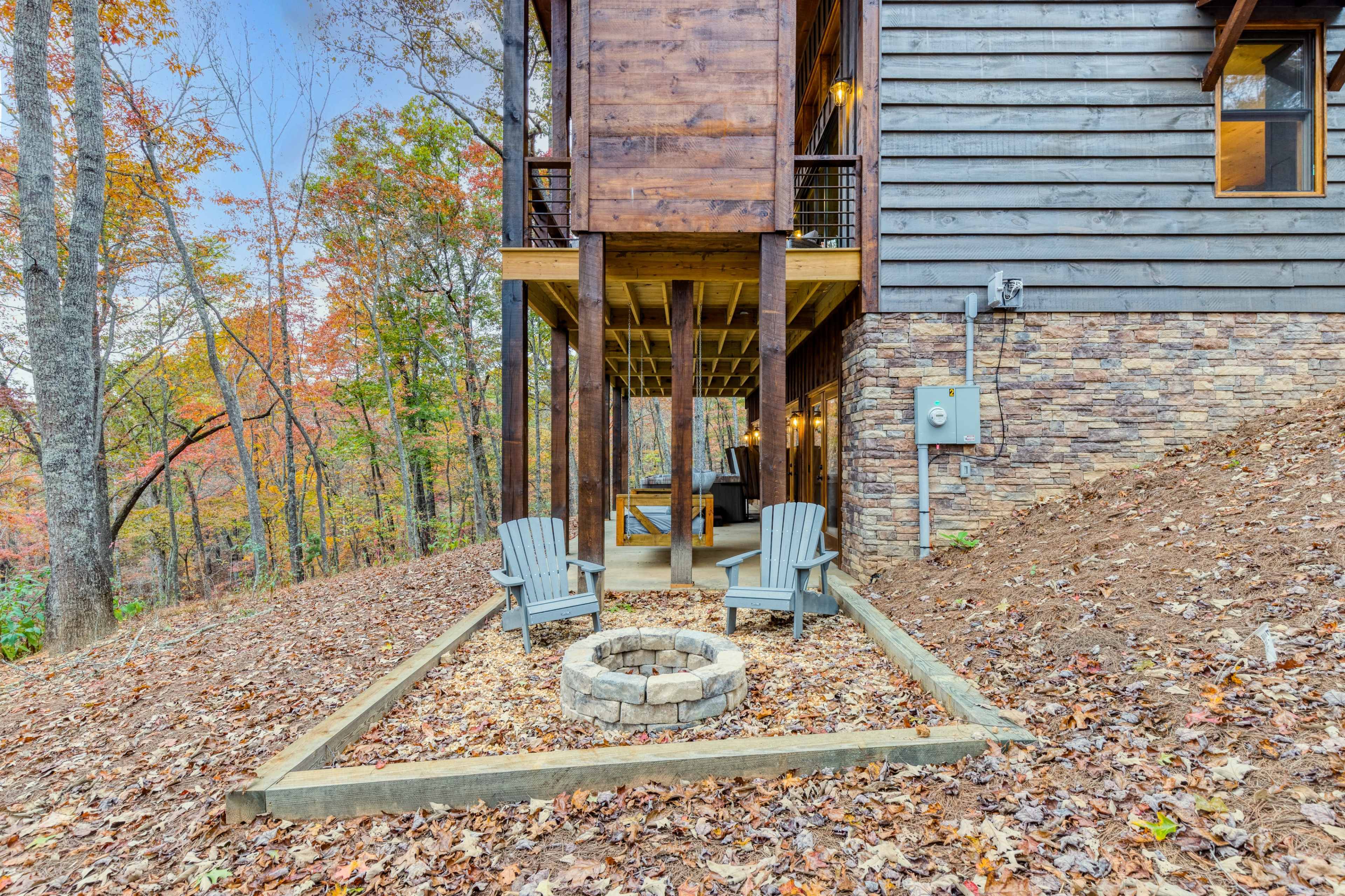 A stone fire pit surrounded by two gray chairs is located beneath a wooden deck of a house, with a backdrop of autumn trees.