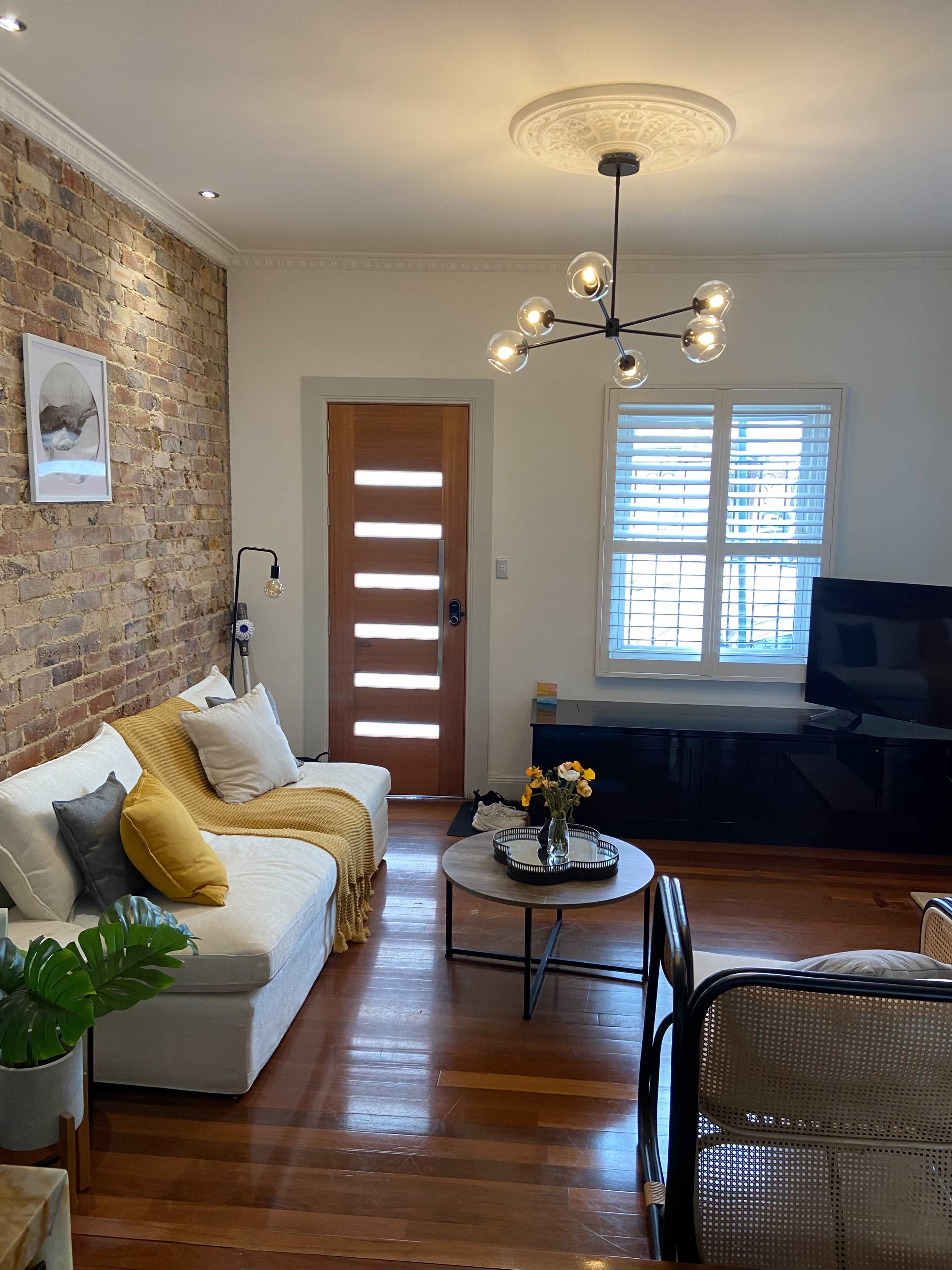 The living room features a brick wall, a white sofa with decorative pillows, a circular coffee table, and a modern light fixture, accented by a wooden door and large windows.