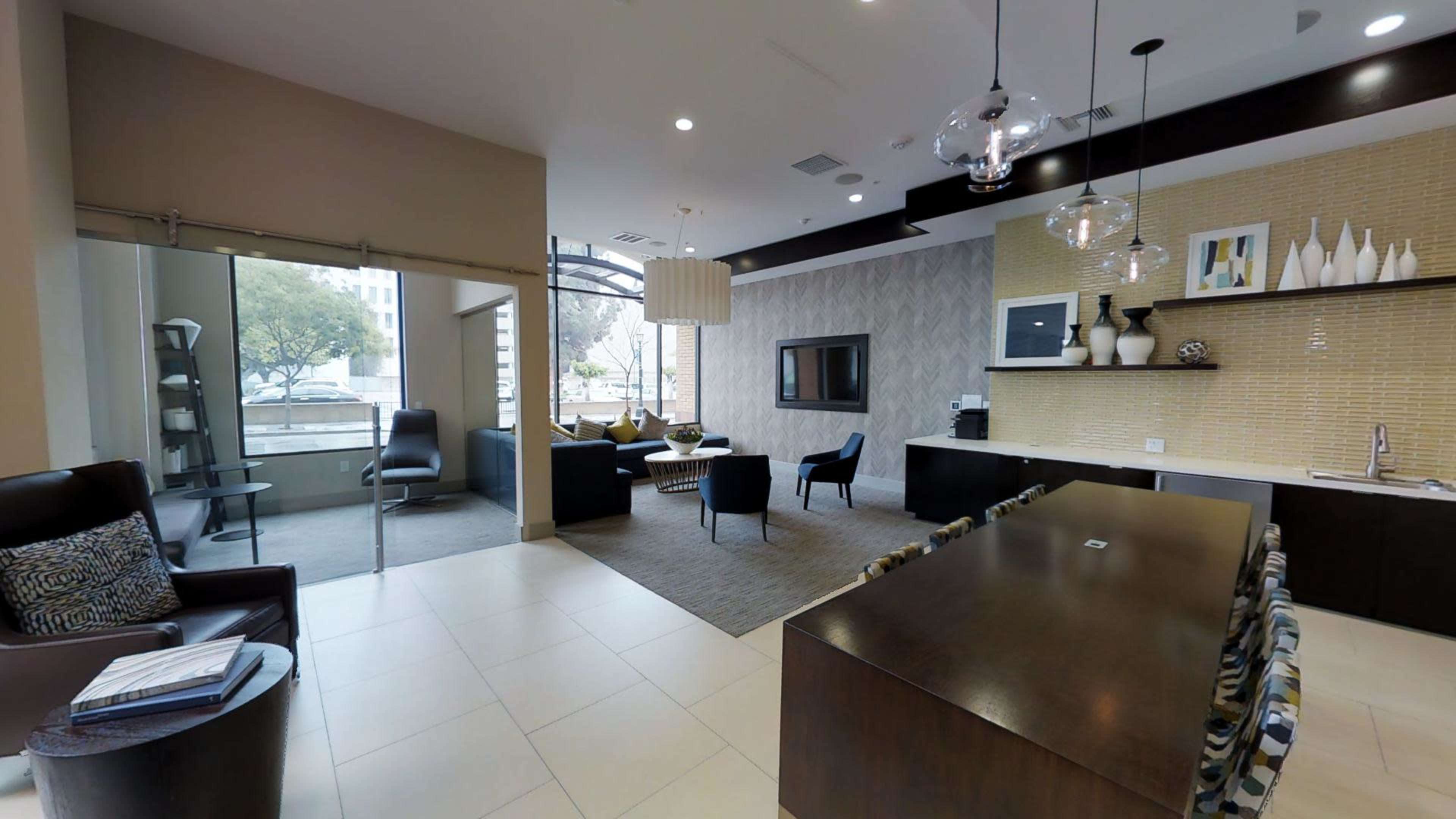 The image shows a modern lounge area featuring a mix of seating arrangements, a kitchenette, and large windows allowing natural light.