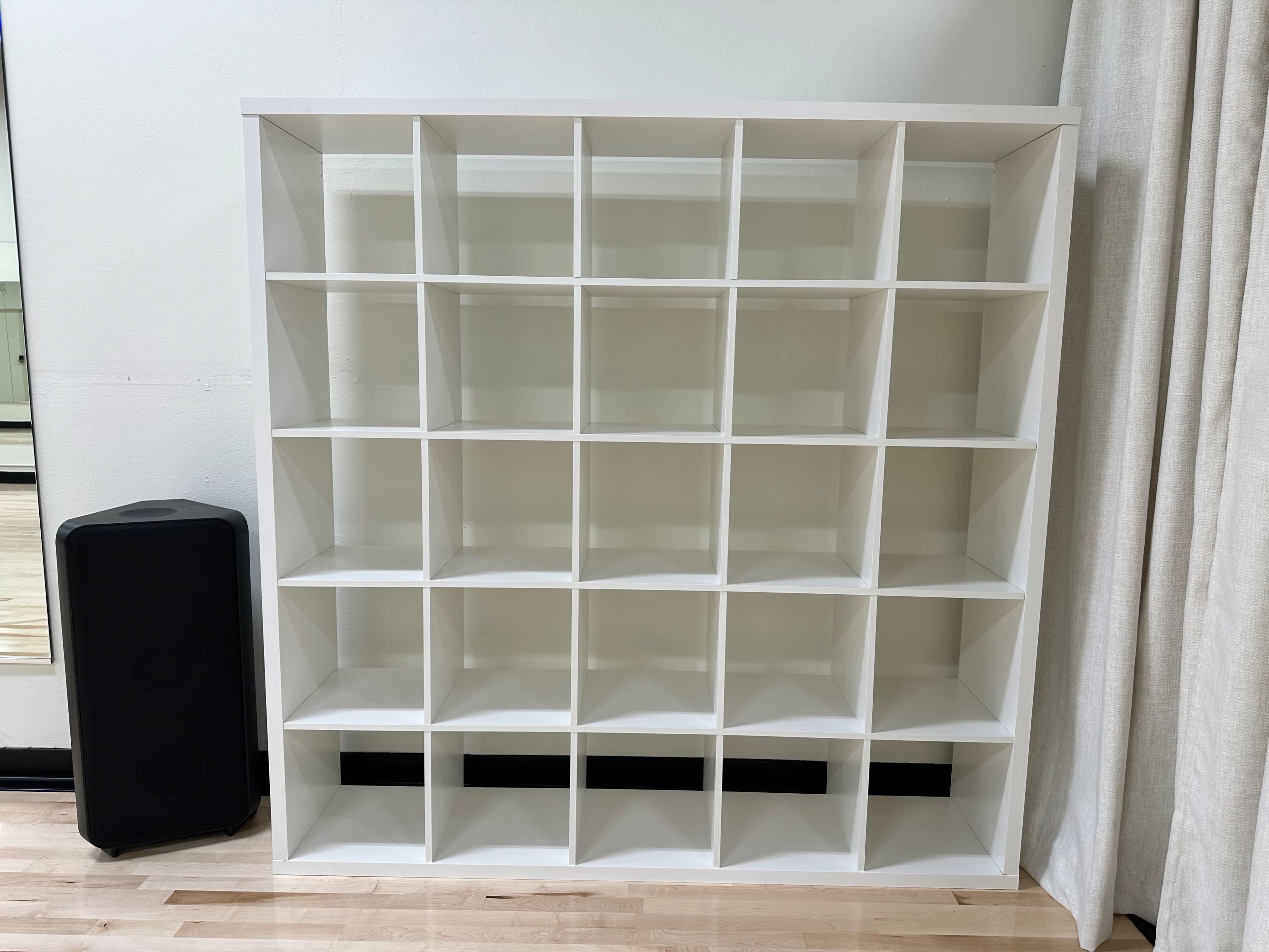 A large white furniture shelving unit with multiple square compartments, positioned next to a black speaker and a curtain in a room.