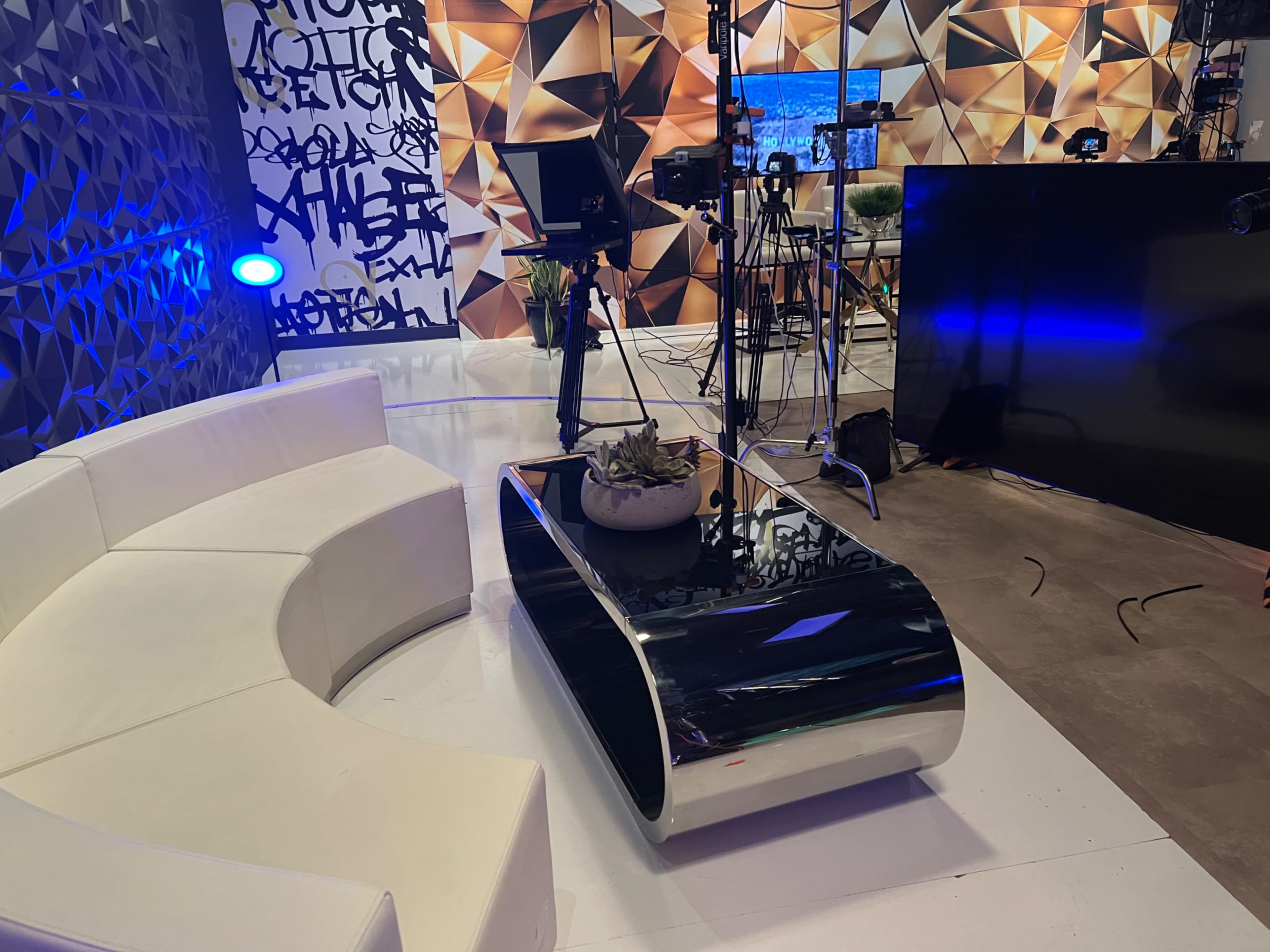 A modern television studio set featuring a white curved sofa, a black shiny coffee table, and multiple cameras on tripods.