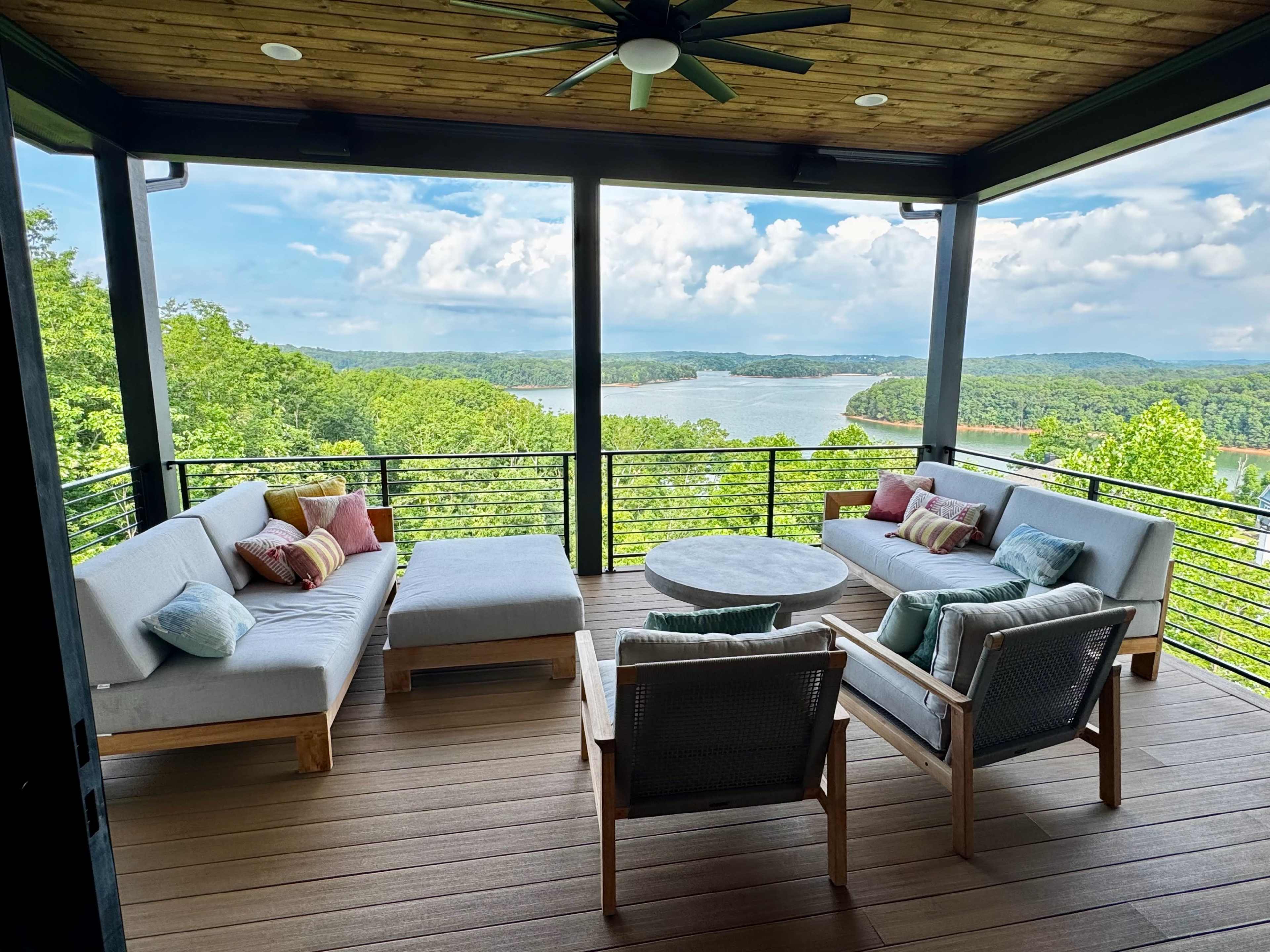 The image shows a spacious terrace with modern seating arrangements overlooking a lake surrounded by lush green hills.