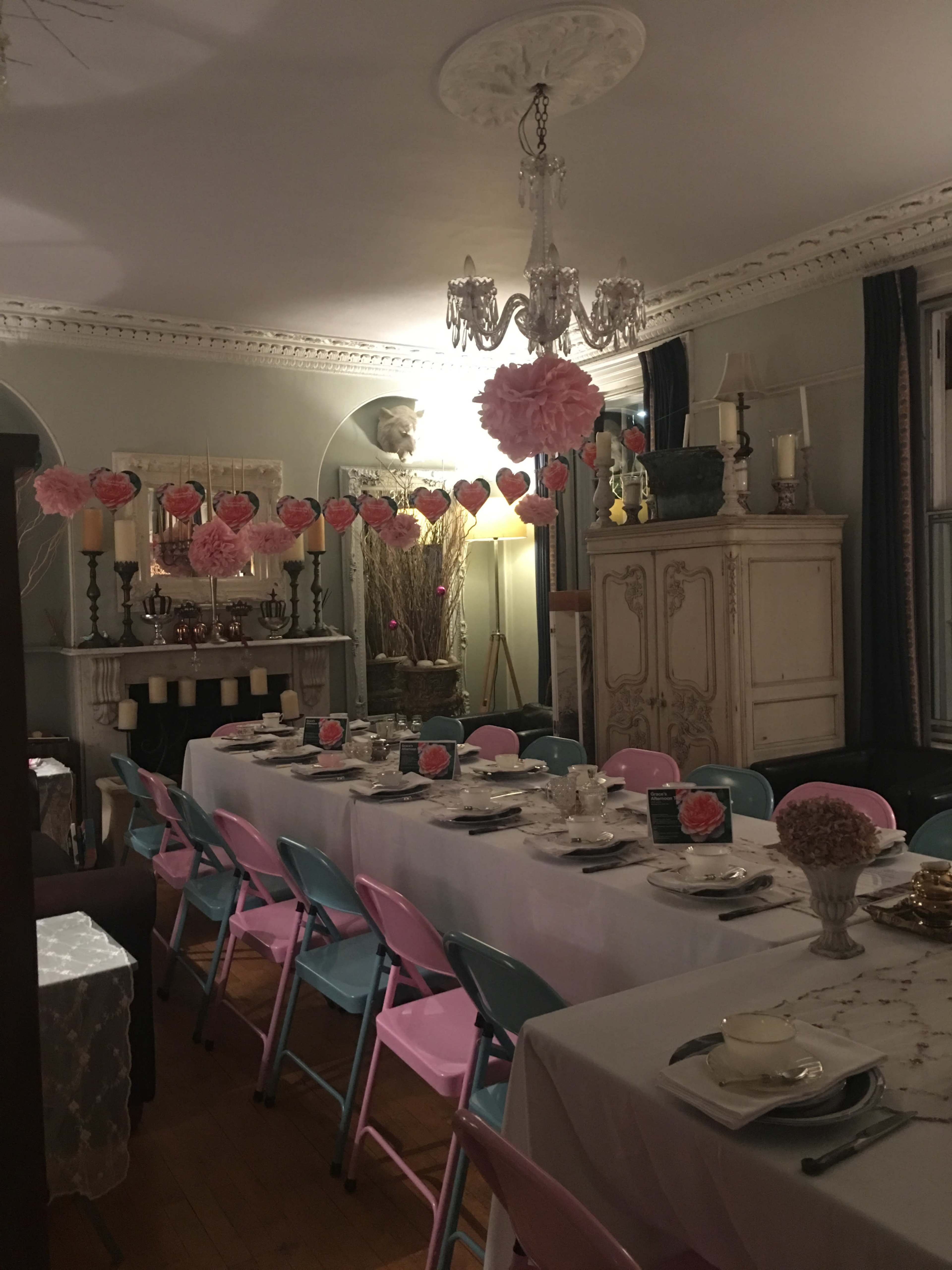 A long dining table is set with white tablecloths and decorative centerpieces, surrounded by pastel-colored chairs in a warmly lit room adorned with hanging paper decorations.