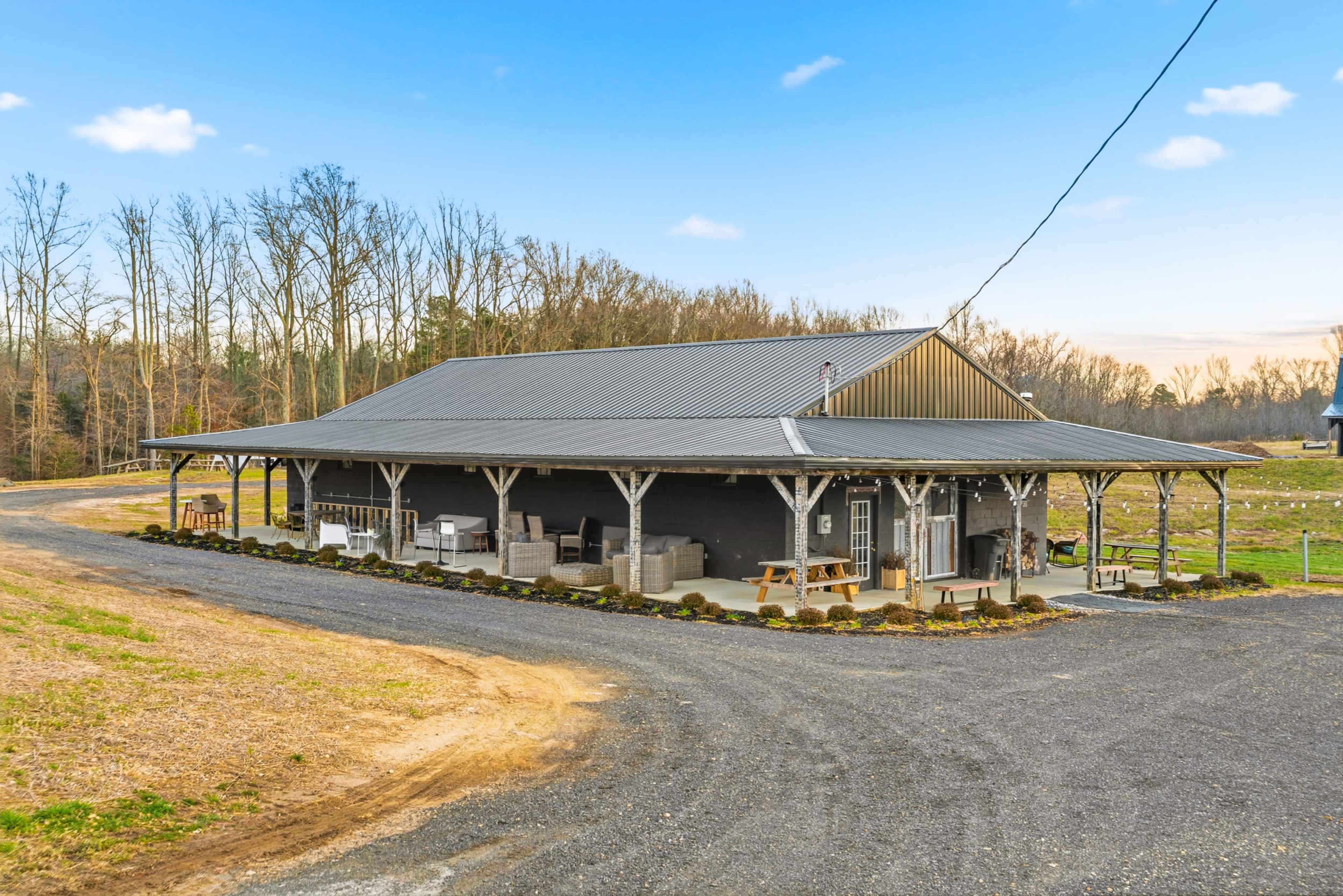 A large, open-sided barn with a metal roof is situated along a gravel path surrounded by trees and open land.