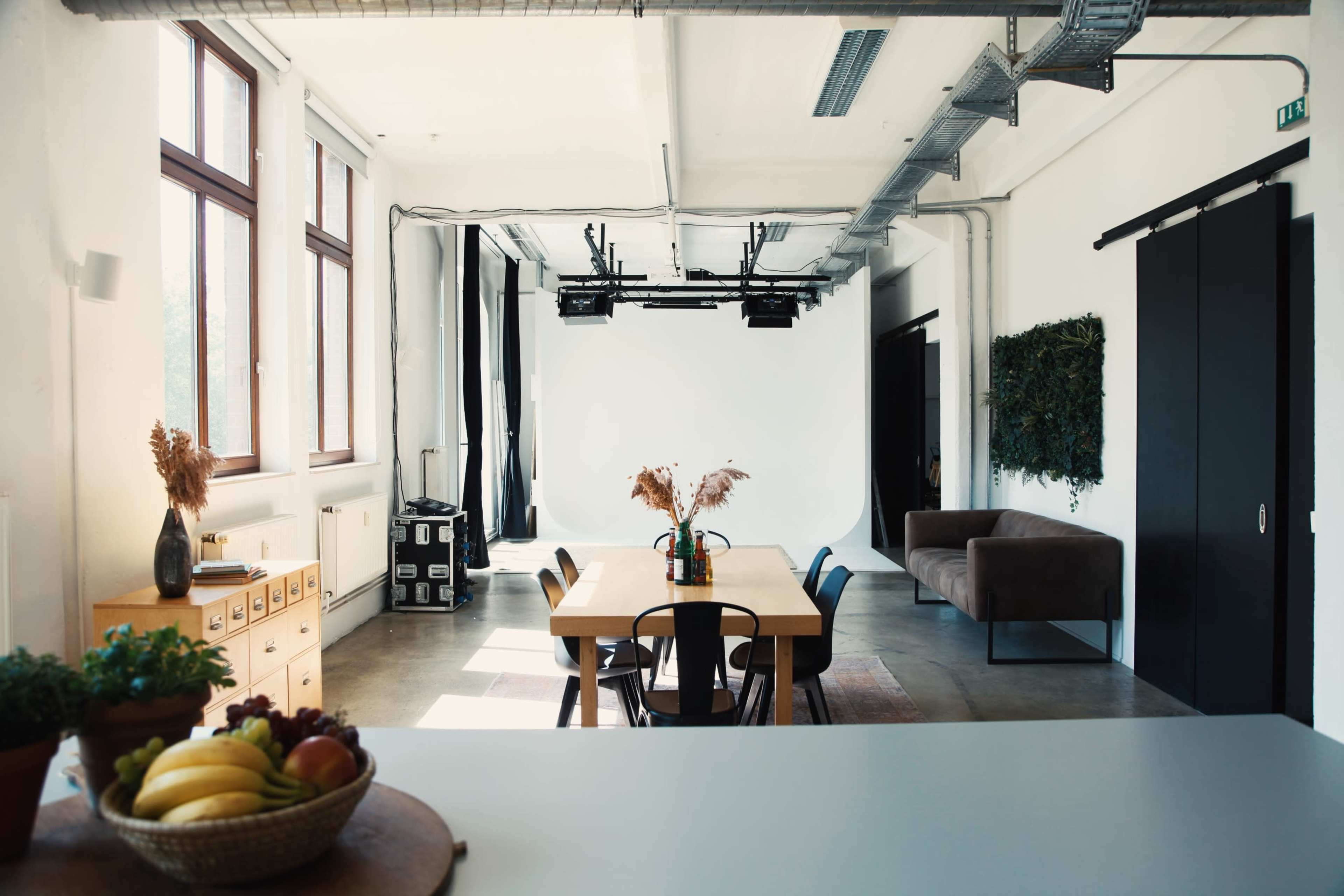 A spacious interior features a dining table with chairs, a bowl of fruit, and a cozy sofa, with large windows allowing natural light to illuminate the space.