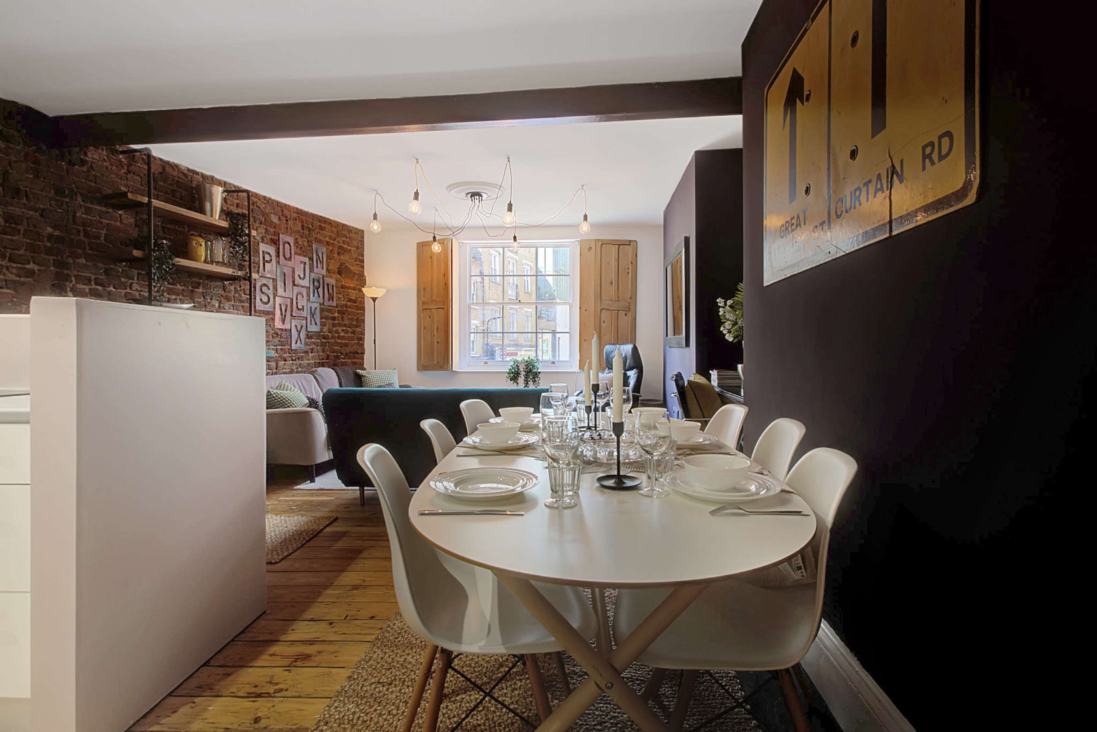 A dining area with a round table and white chairs is situated next to a living space featuring exposed brick walls and large windows.