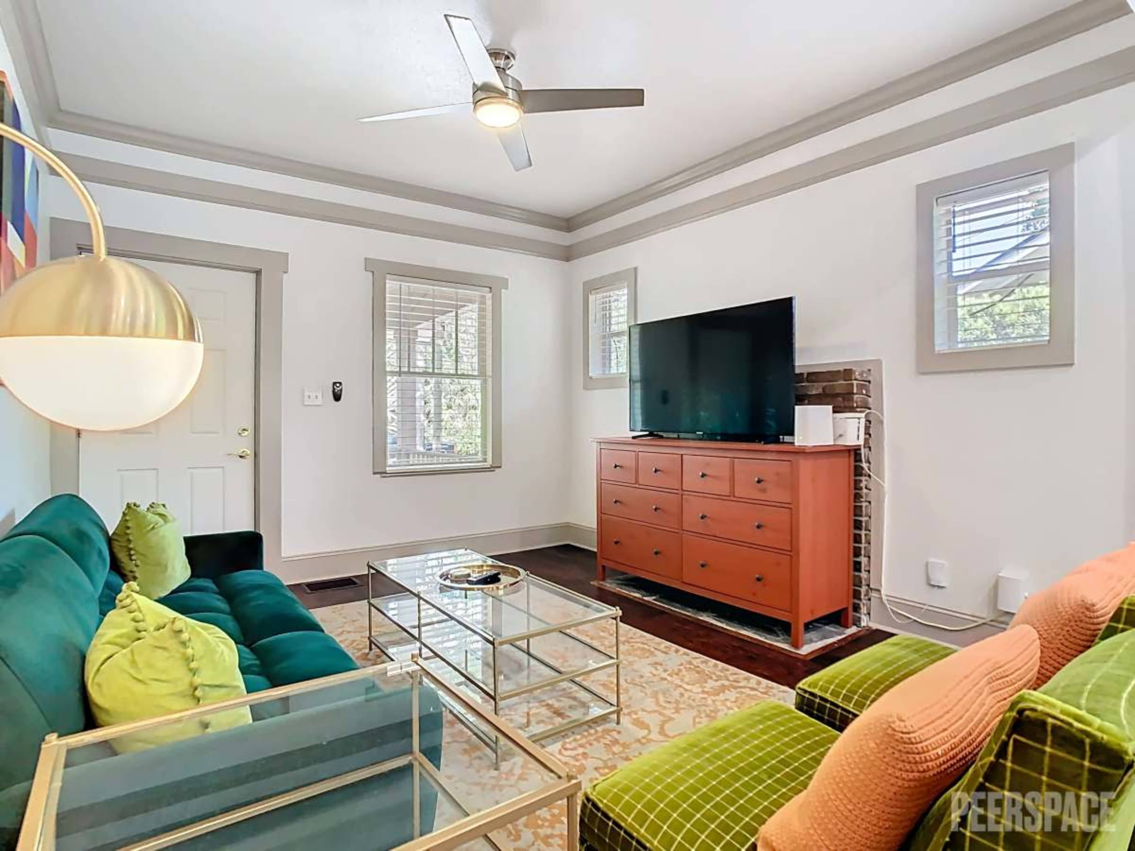 A cozy living room features a green sofa, an orange accent chair, a glass coffee table, and a large TV mounted on the wall.