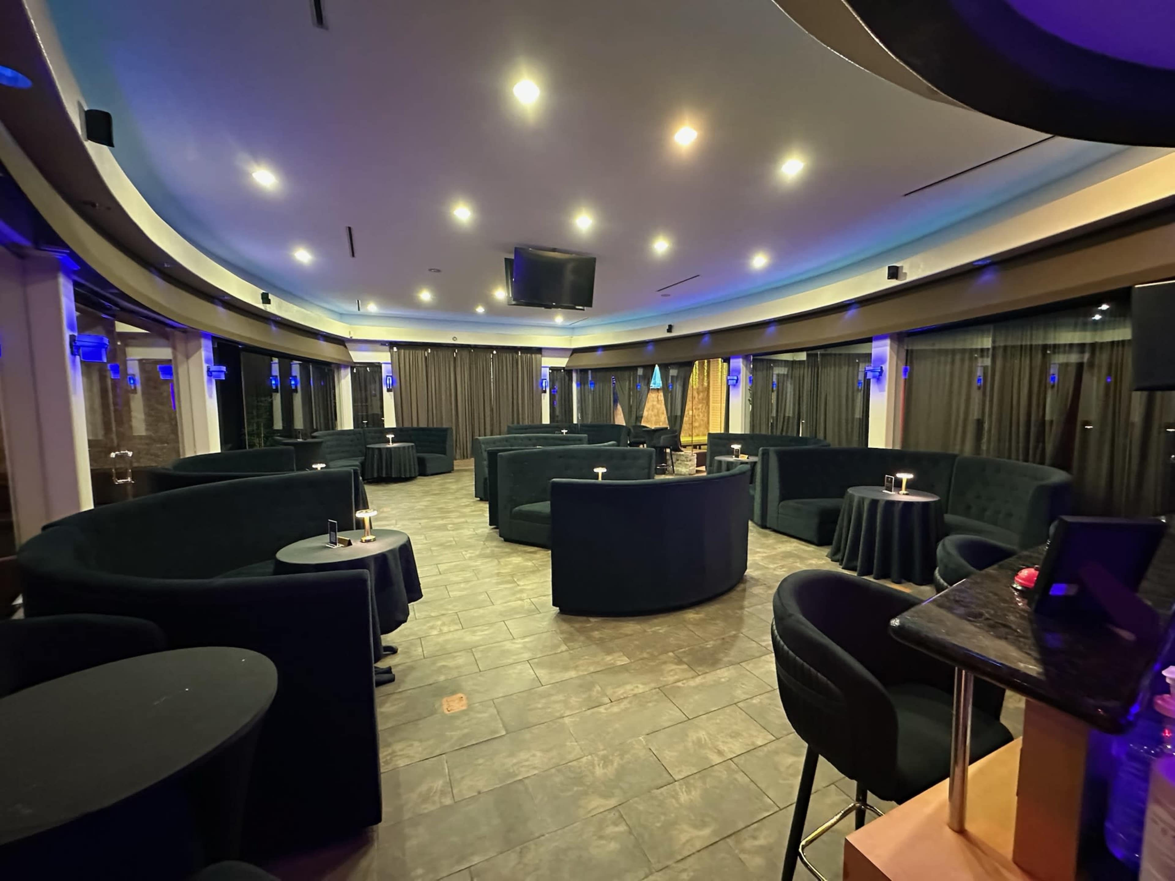 The image shows a spacious lounge area with black seating, small round tables, and dim lighting.