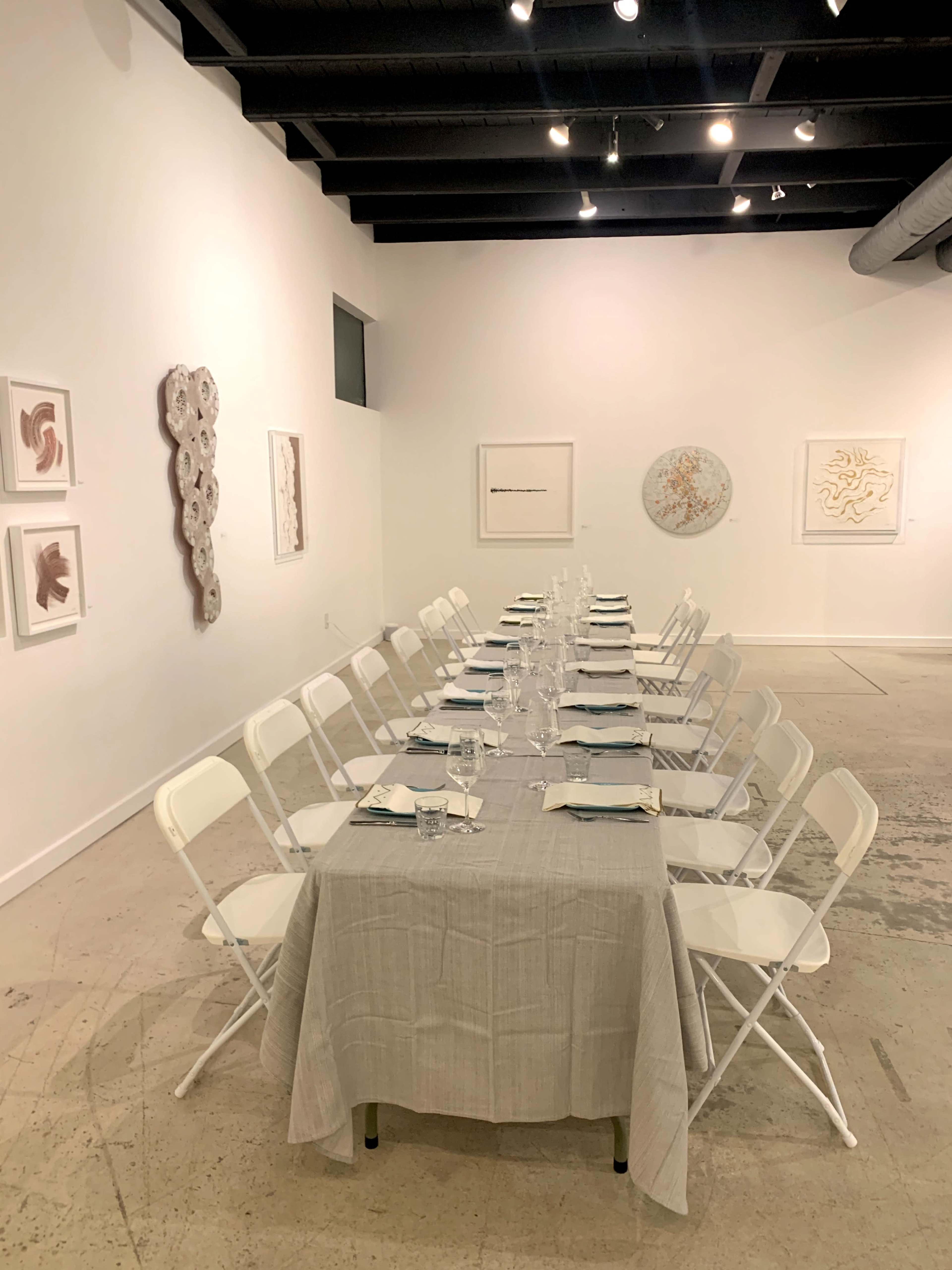 A long table is set with white chairs and glassware in an art gallery featuring light-colored walls and various artworks.