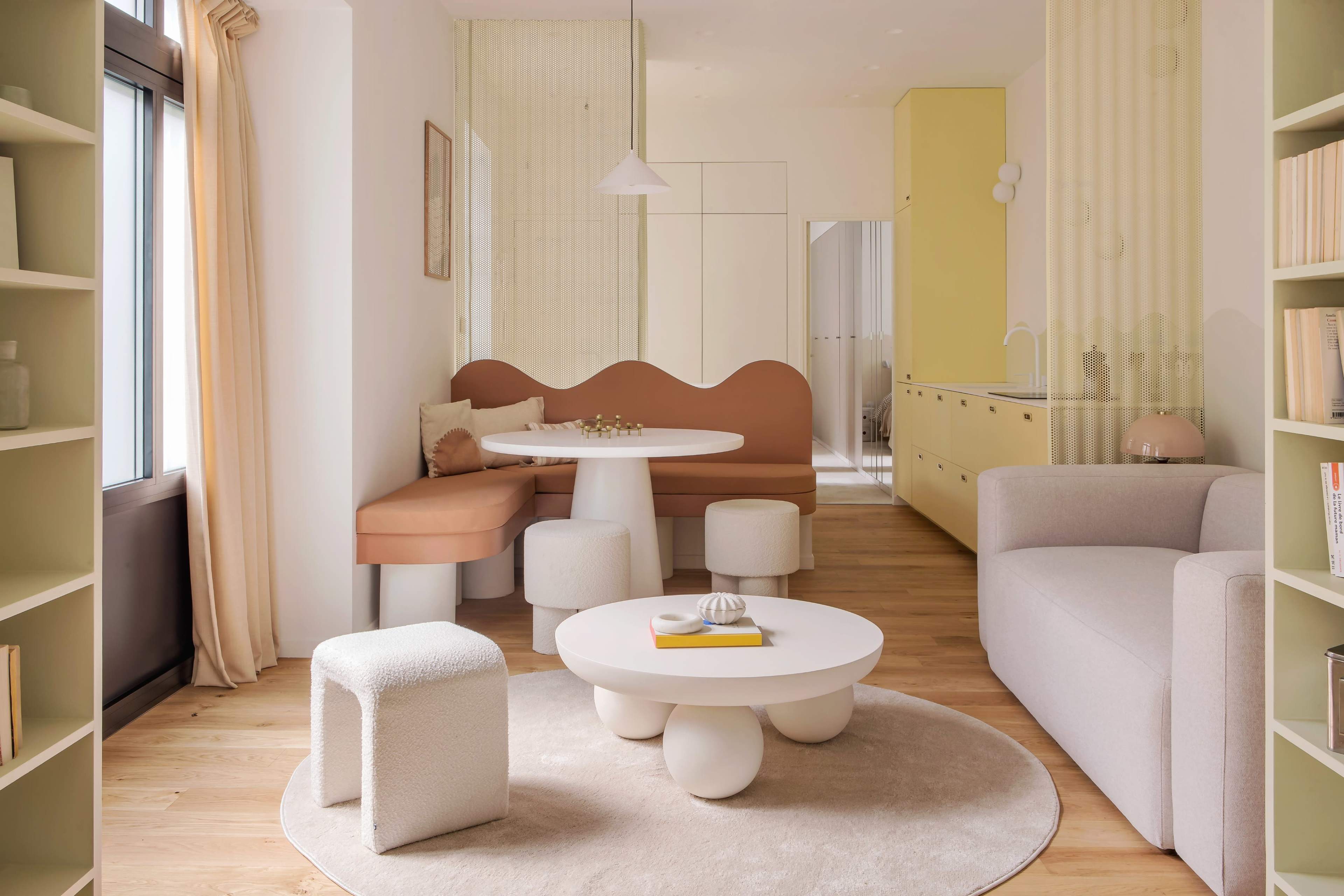 The image shows a modern, minimalist living space featuring a round dining table surrounded by soft seating, a light color palette, and decorative elements like a circular coffee table and pastel-colored walls.
