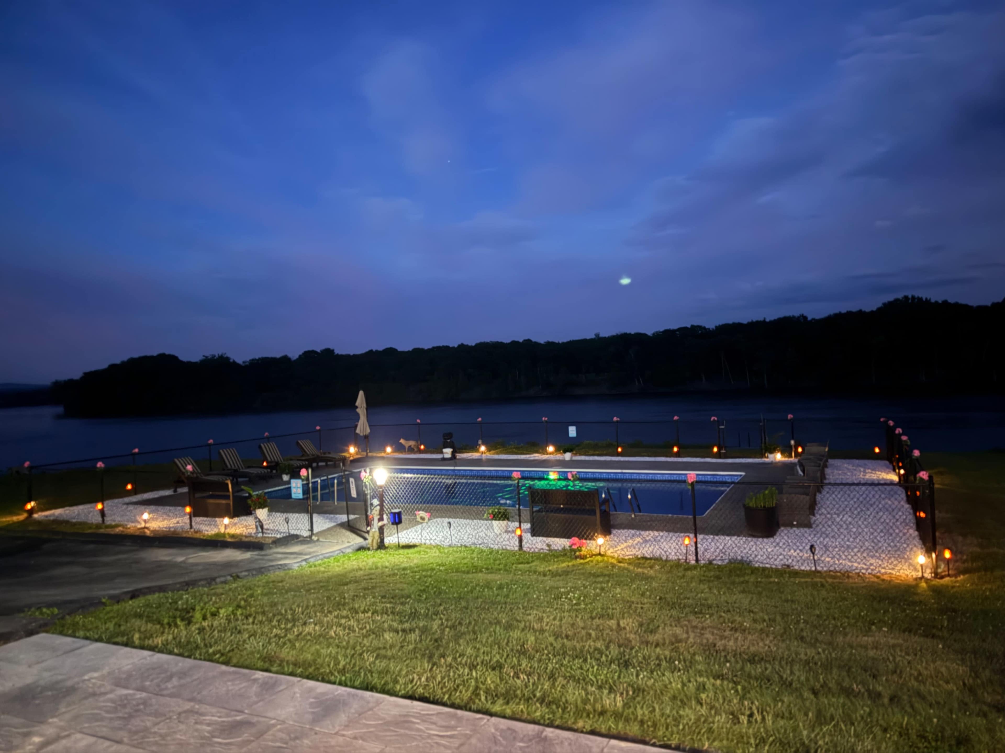 A swimming pool surrounded by lights and a grassy area is visible near a body of water at dusk.
