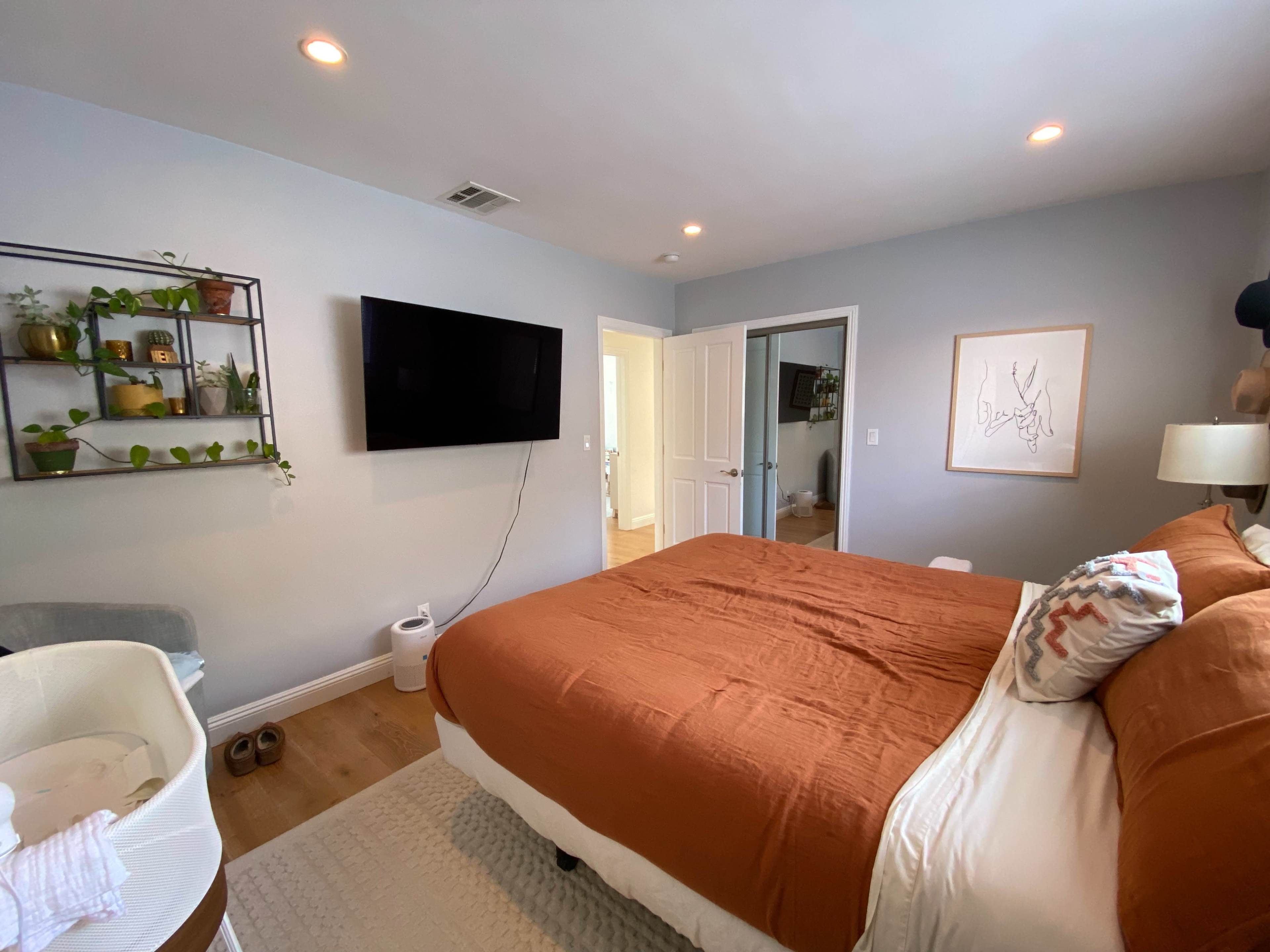 A well-lit bedroom features a neatly made bed with an orange duvet, a wall-mounted TV, and a plant shelf.