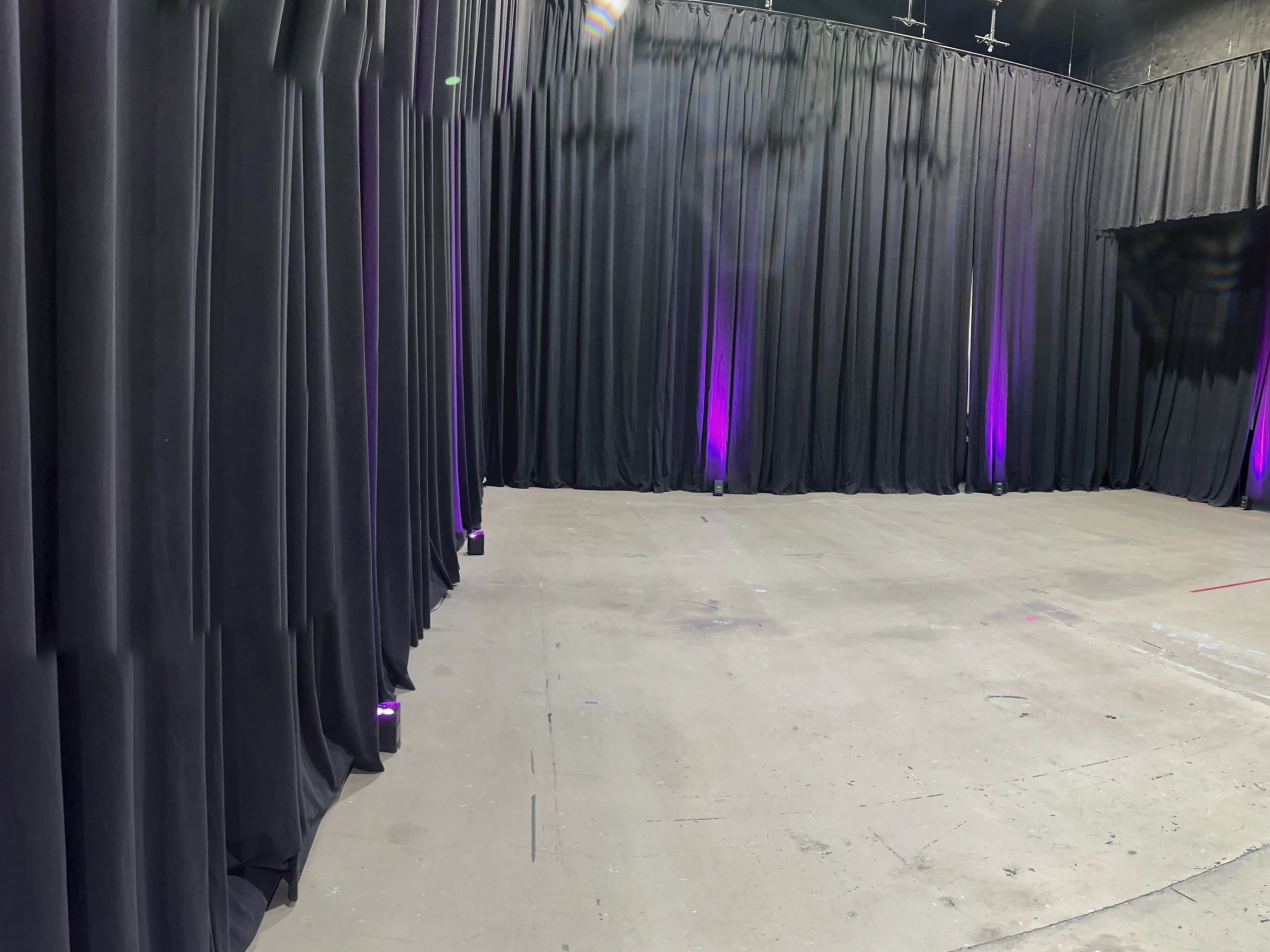 A large empty space with black curtains lining the walls and purple lights illuminating the floor.