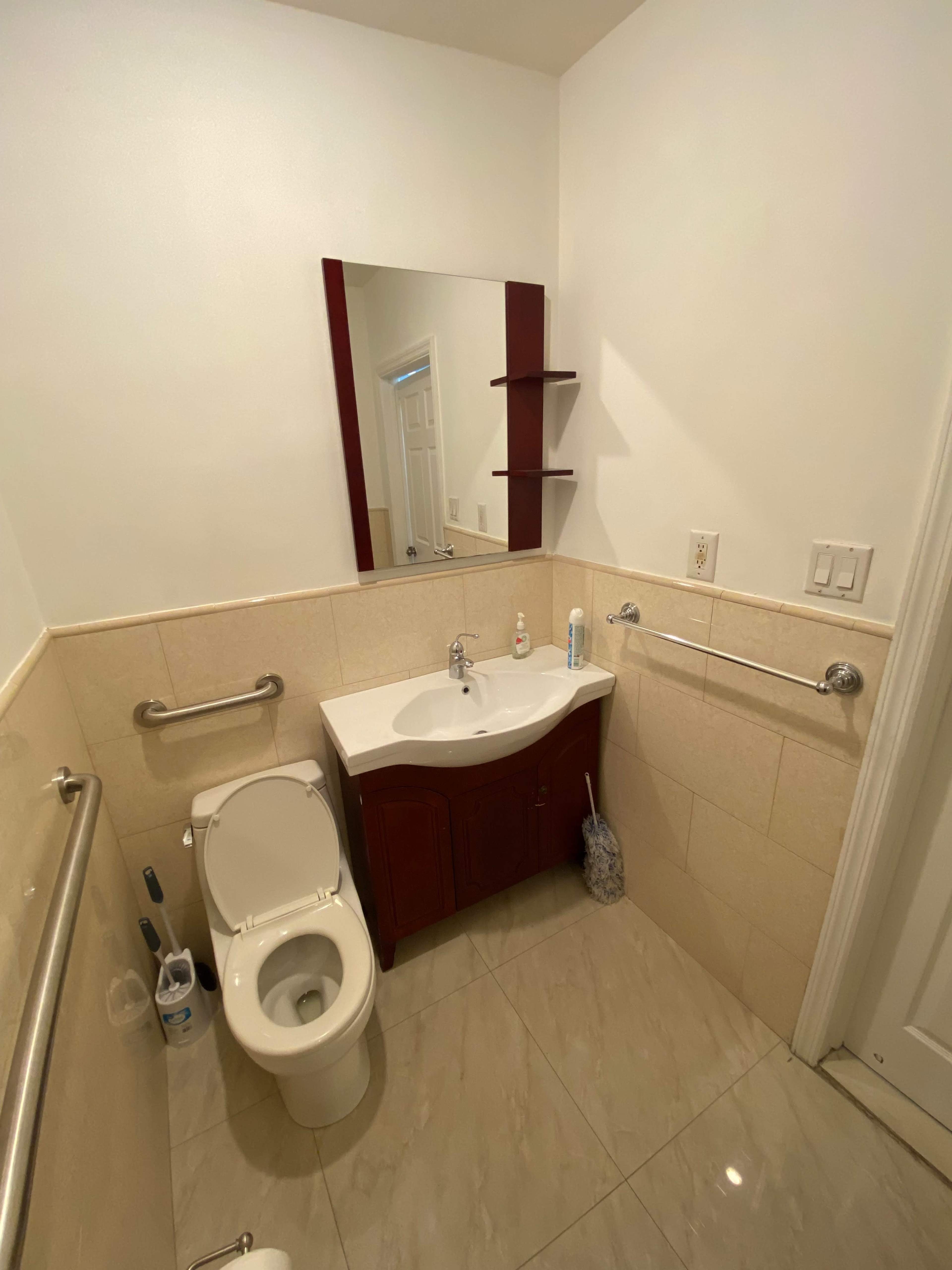 A small, well-lit bathroom featuring a toilet, sink with a cabinet underneath, a mirror, and a few shelves on the wall.