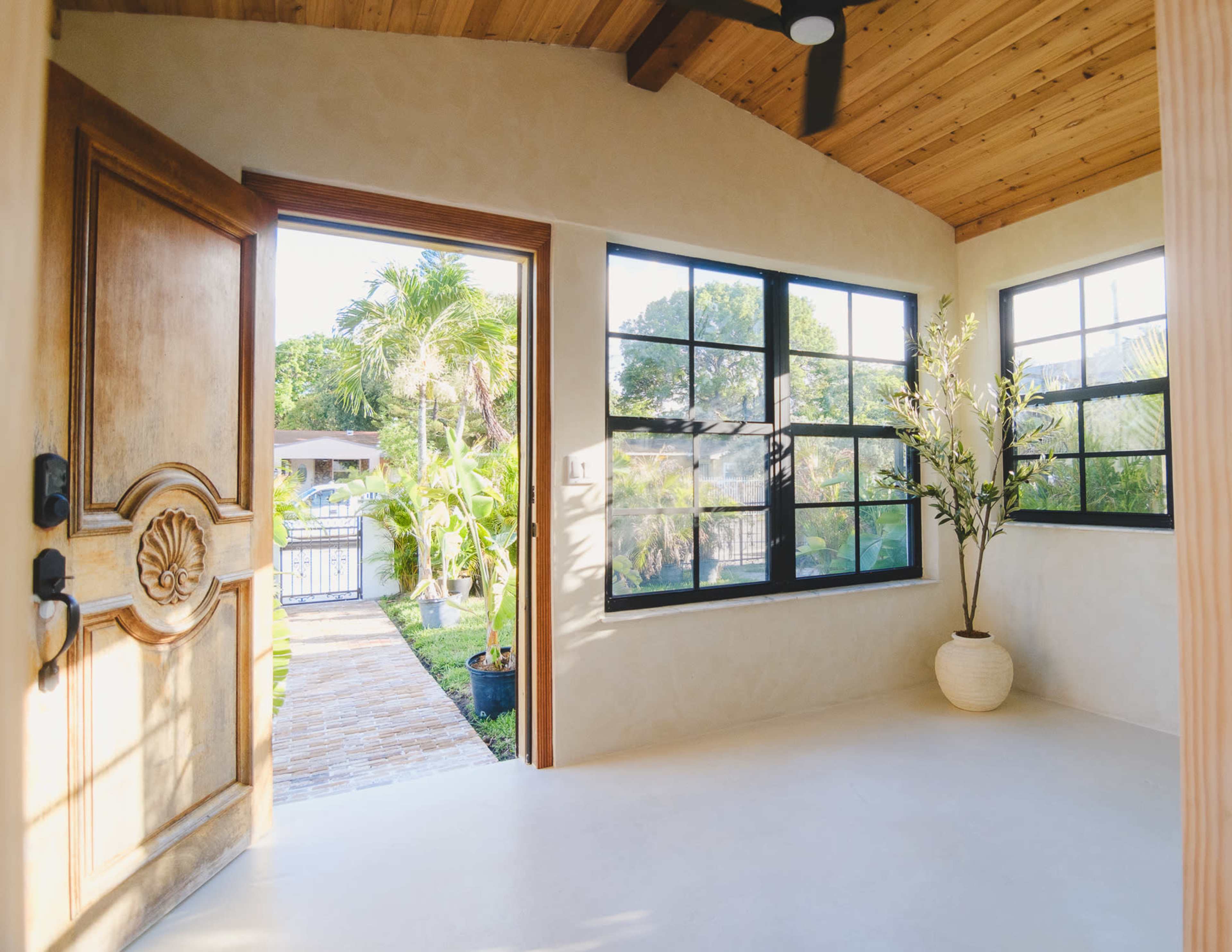 A bright interior features an open door leading to a path outside, framed by large windows and a simple potted plant.