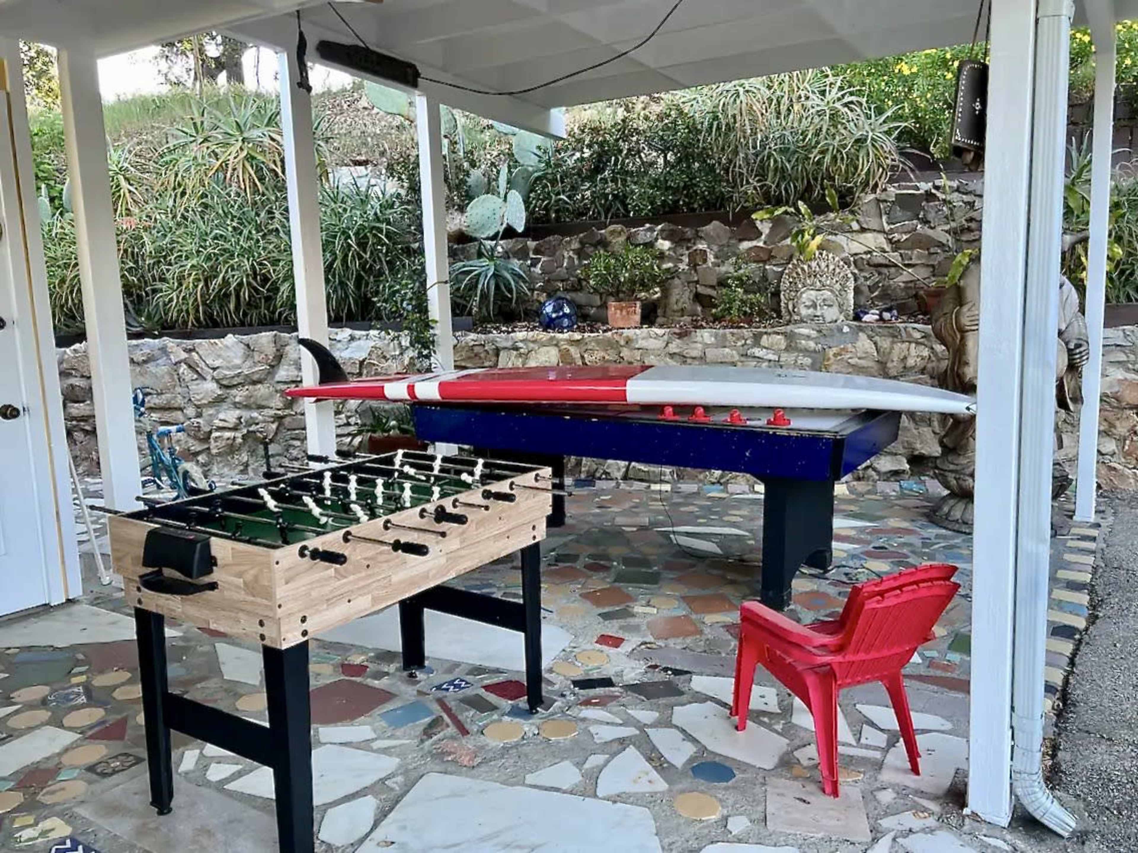 An outdoor game area featuring a foosball table, a pool table with cues, and a red plastic chair under a covered patio surrounded by greenery.