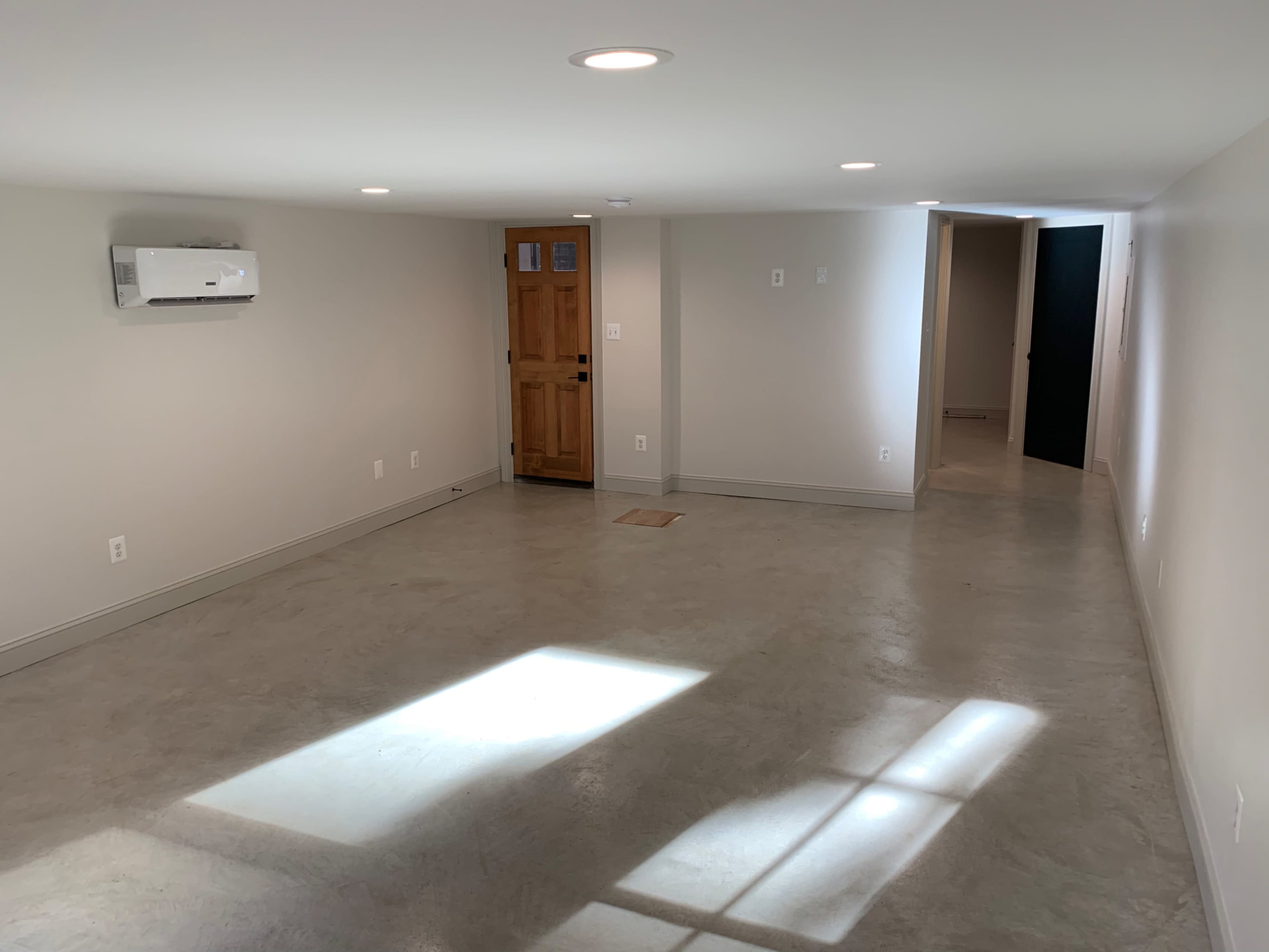A spacious, well-lit basement with a concrete floor, an air conditioning unit, a doorway to the outside, and a dark door leading to another room.