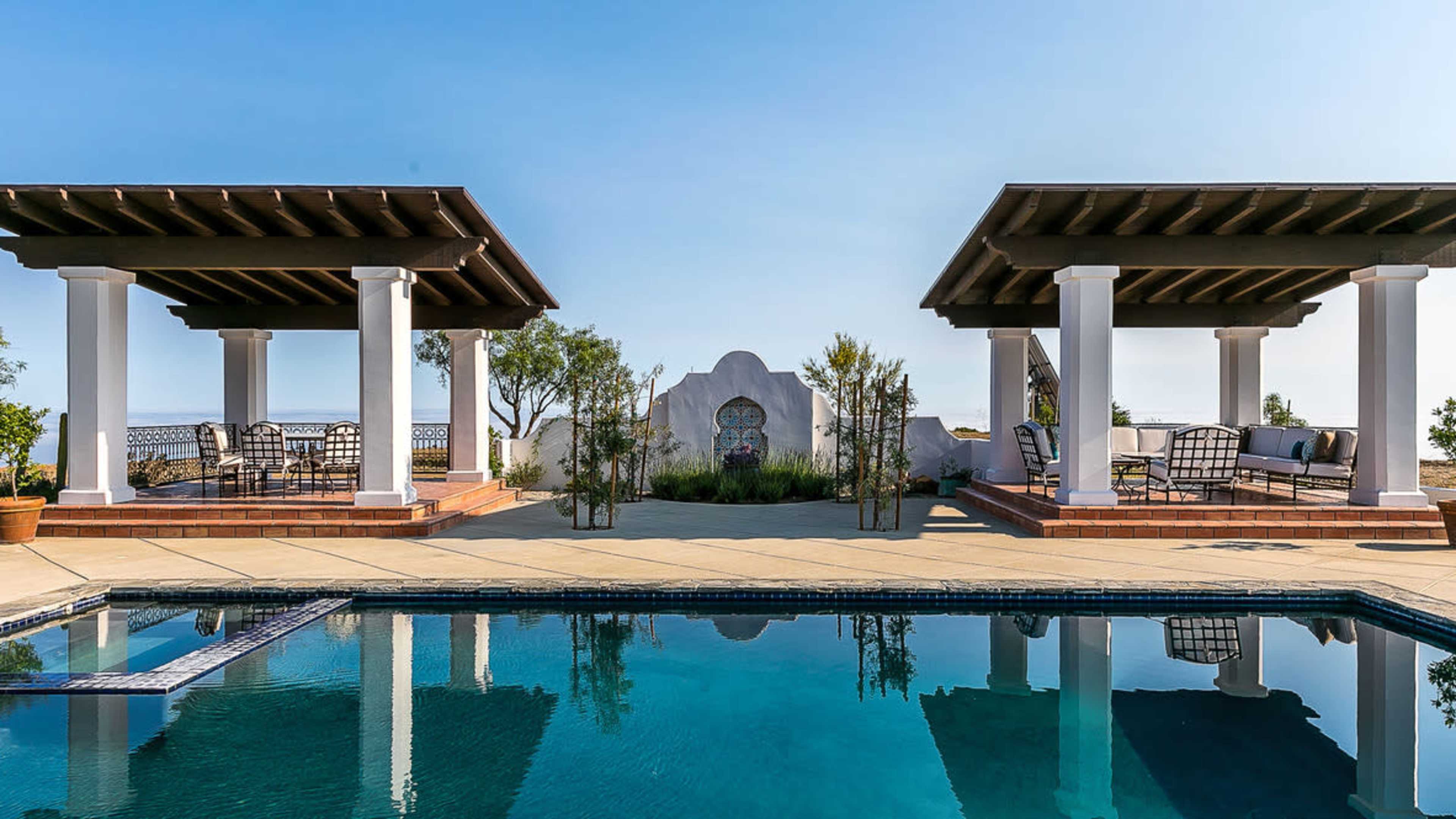 A landscaped pool area with two covered seating areas on either side and a decorative wall in the background.