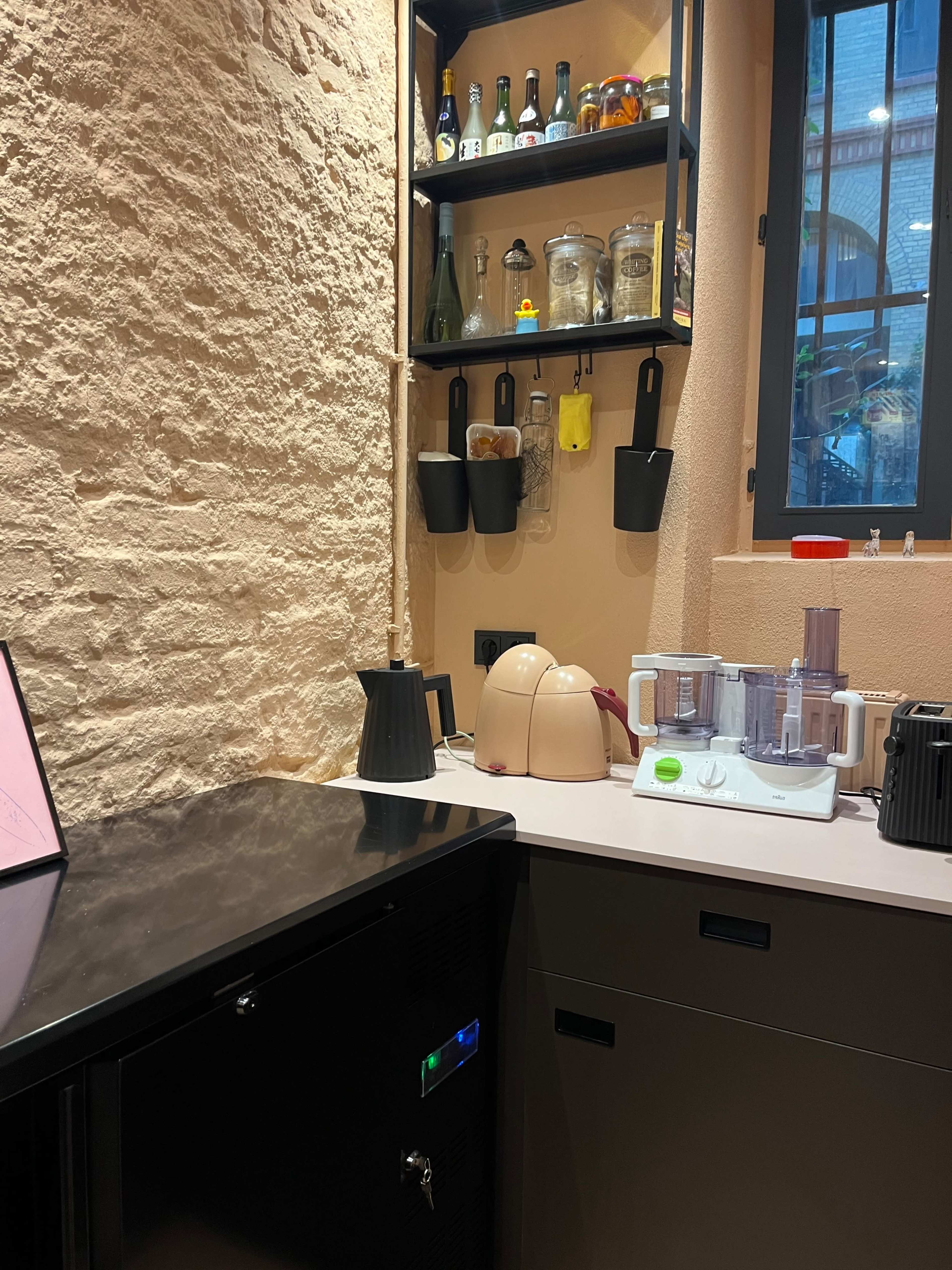 The image shows a kitchen corner with a black countertop, an under-counter fridge, a food processor, and a shelf holding various jars and bottles.