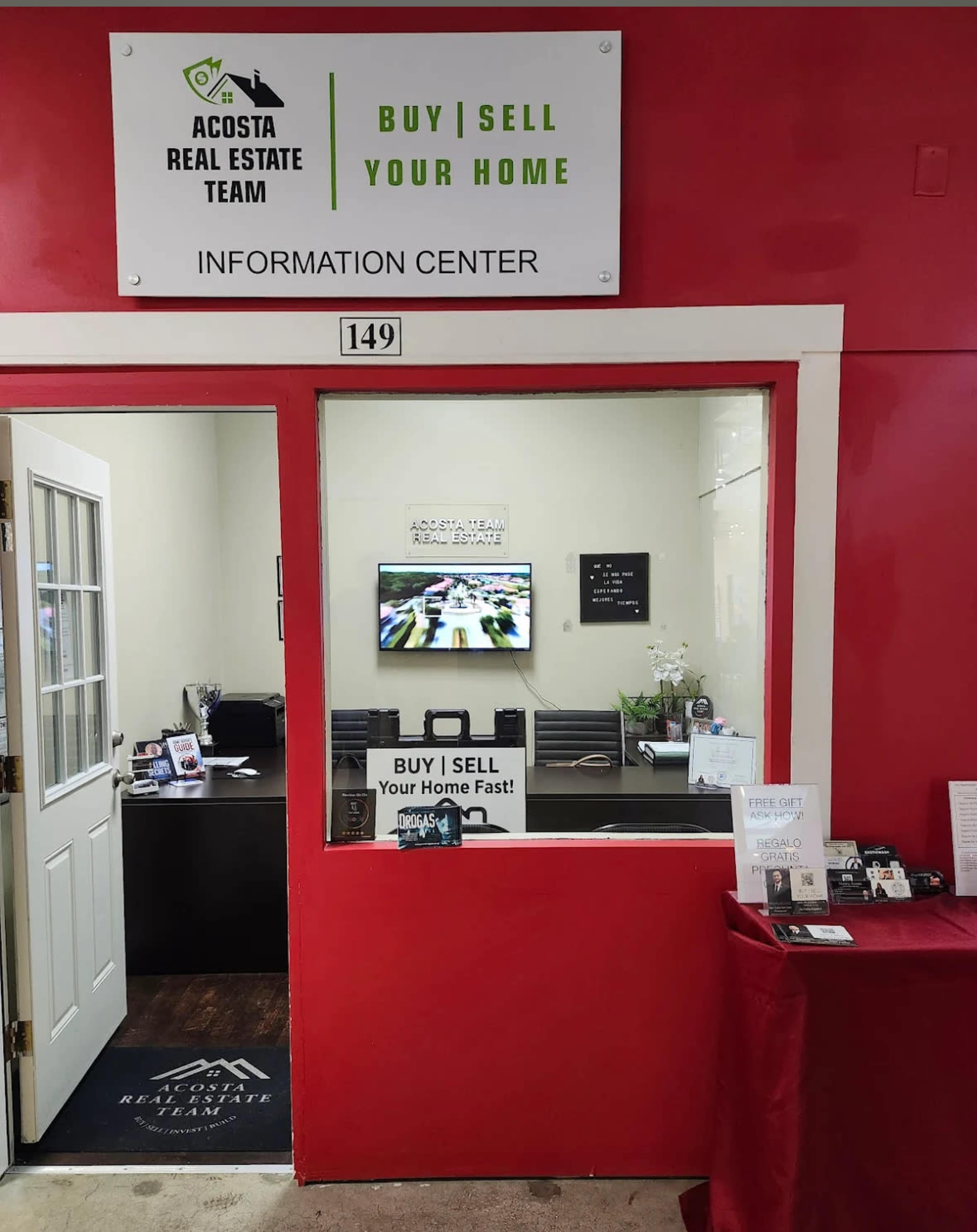 The image shows a real estate information center with a red wall, a desk visible through an open door, and a sign above stating "ACOSTA REAL ESTATE TEAM" and "BUY | SELL | YOUR HOME."