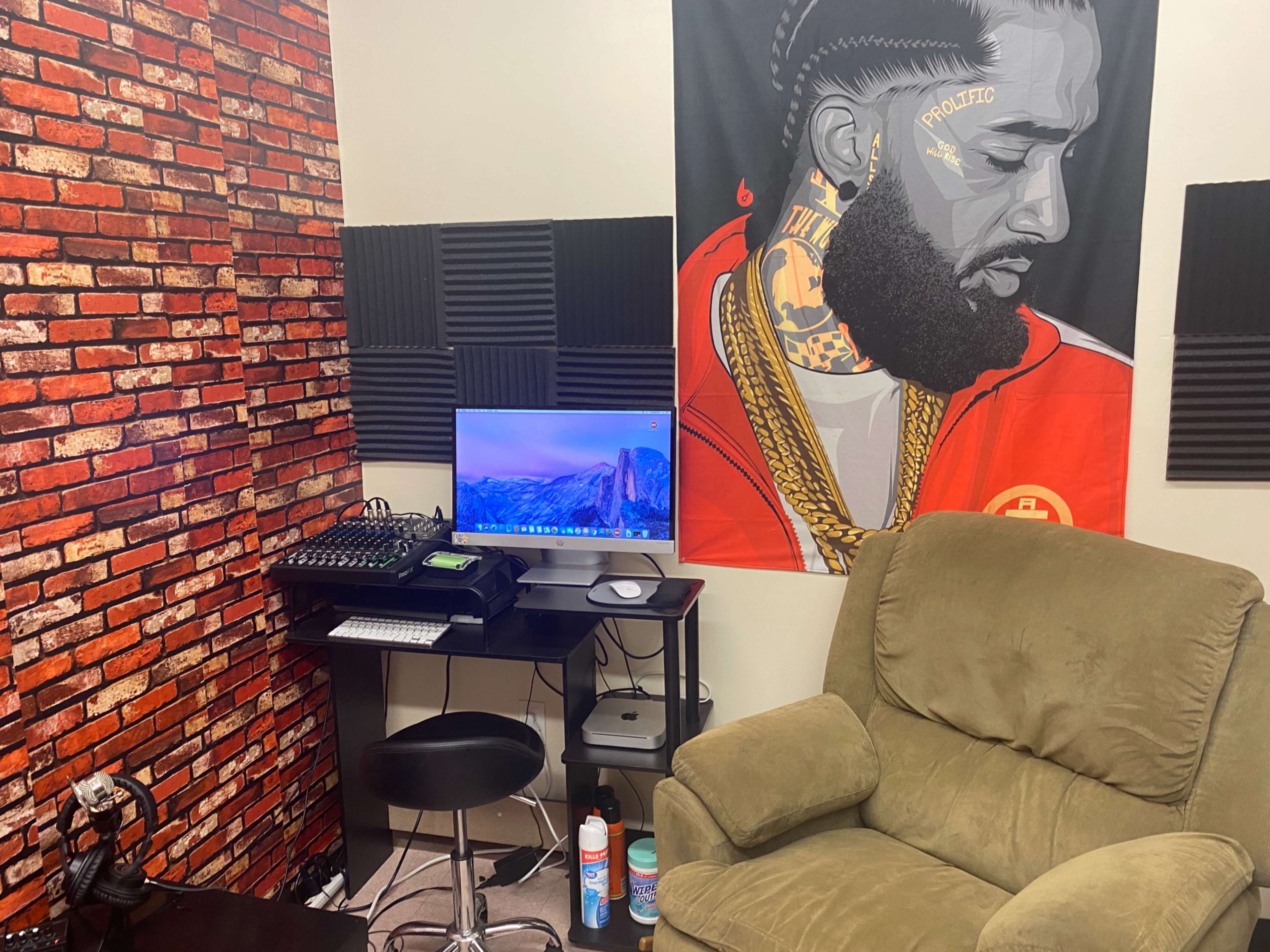 A small room featuring a desk with a computer monitor, a green armchair, and a wall adorned with a mural of a man and a brick pattern.