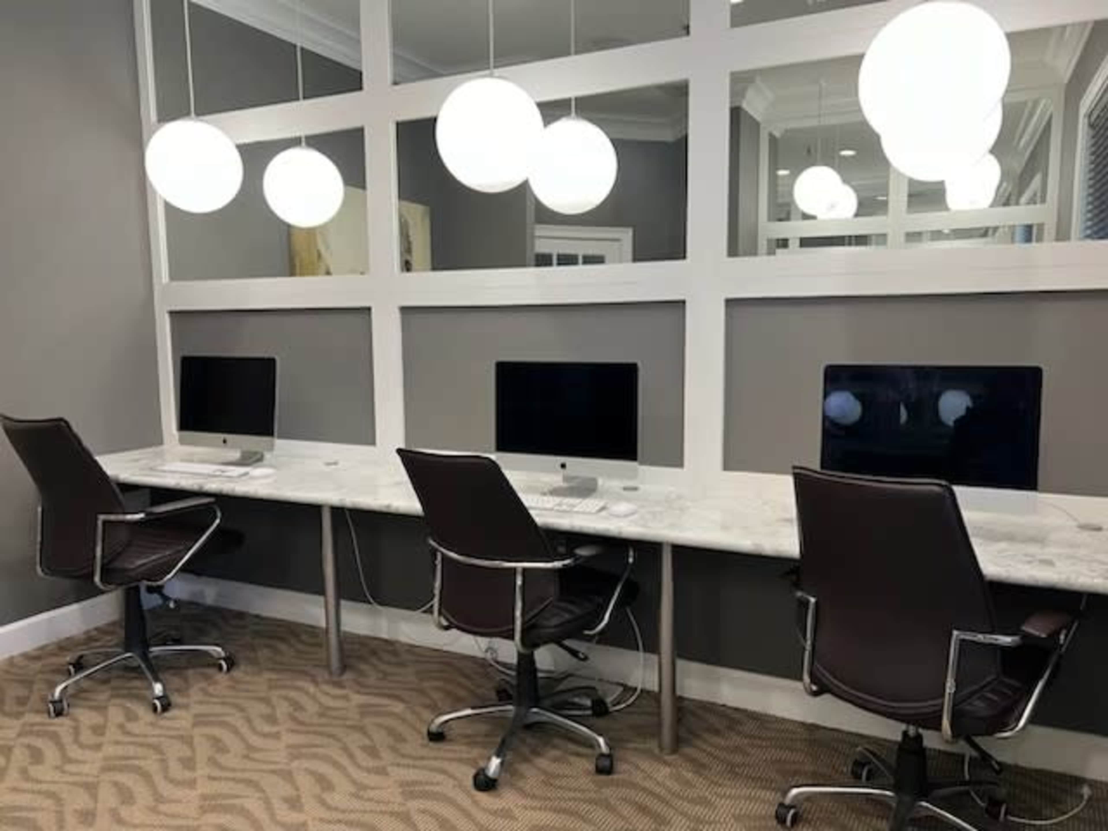 A modern workspace with three desktop computers on marble-top desks, each accompanied by swivel chairs and illuminated by round pendant lights above.