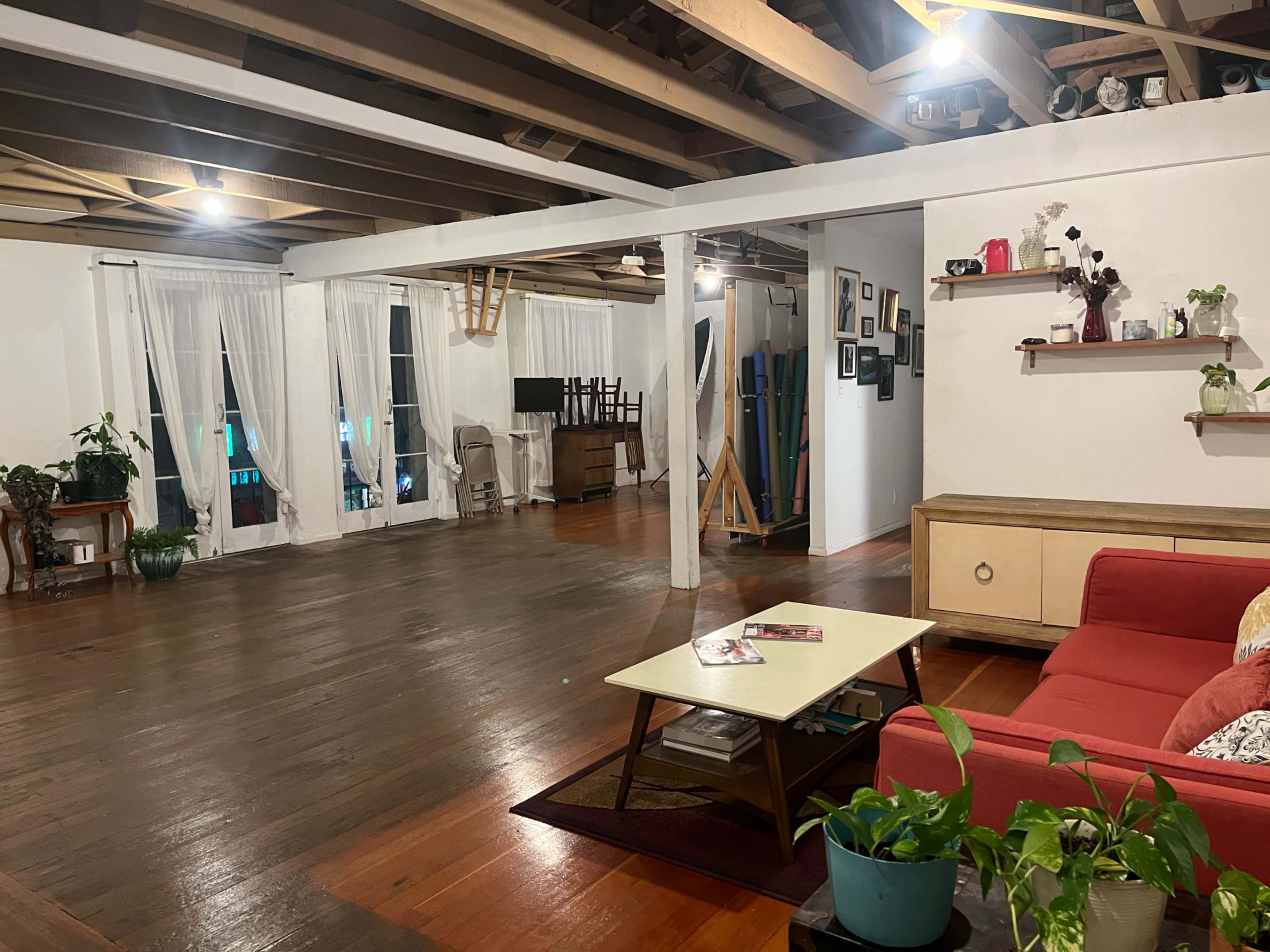 A spacious, well-lit living area with wooden floors, large windows covered by white curtains, a red couch, and various pieces of furniture and plants.