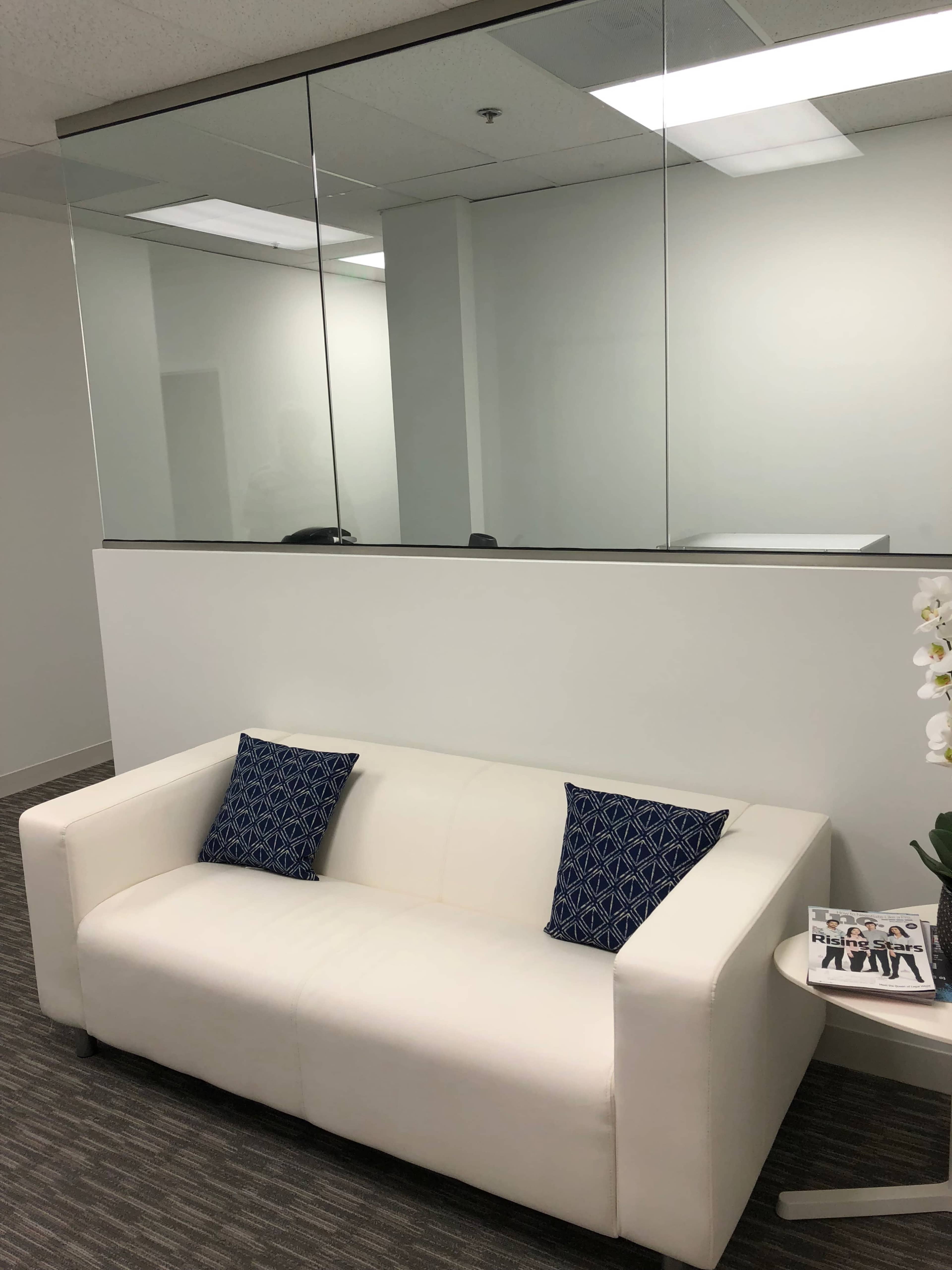 A white sofa with two blue patterned pillows is positioned against a white wall in a well-lit office space featuring a glass partition.