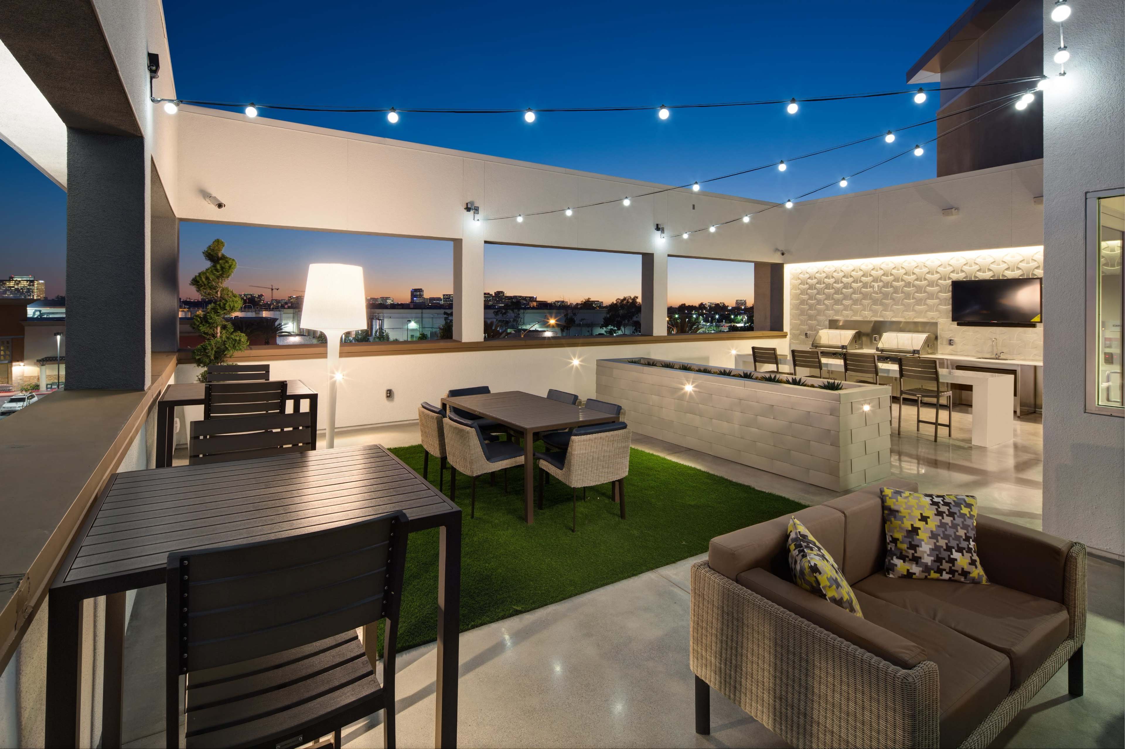 The image shows a modern rooftop patio featuring several seating areas, outdoor furniture, and string lights illuminating the space against a twilight sky.