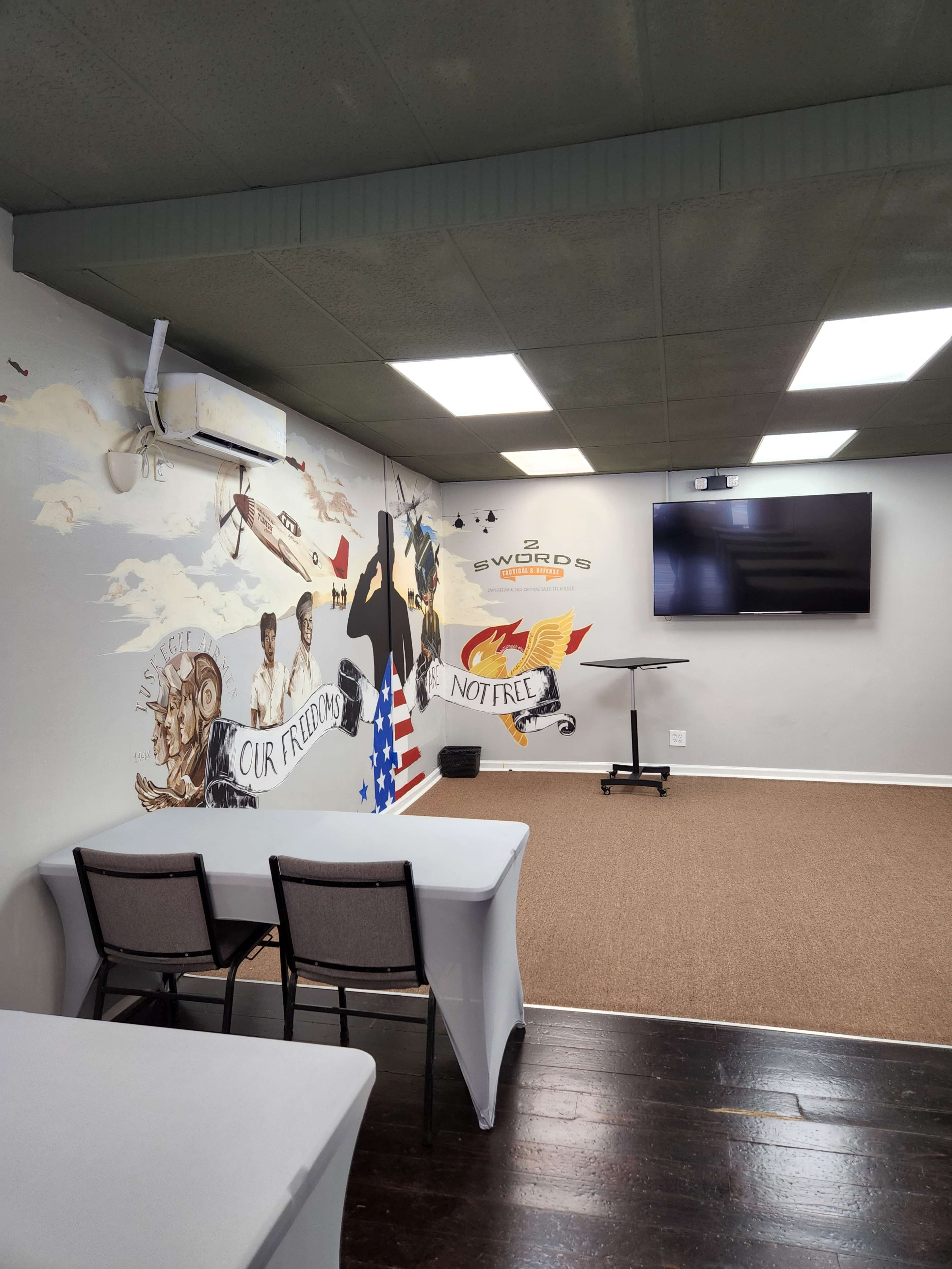 The room features a mural on one wall with patriotic imagery, two empty chairs in front of tables, and a television mounted on the opposite wall.