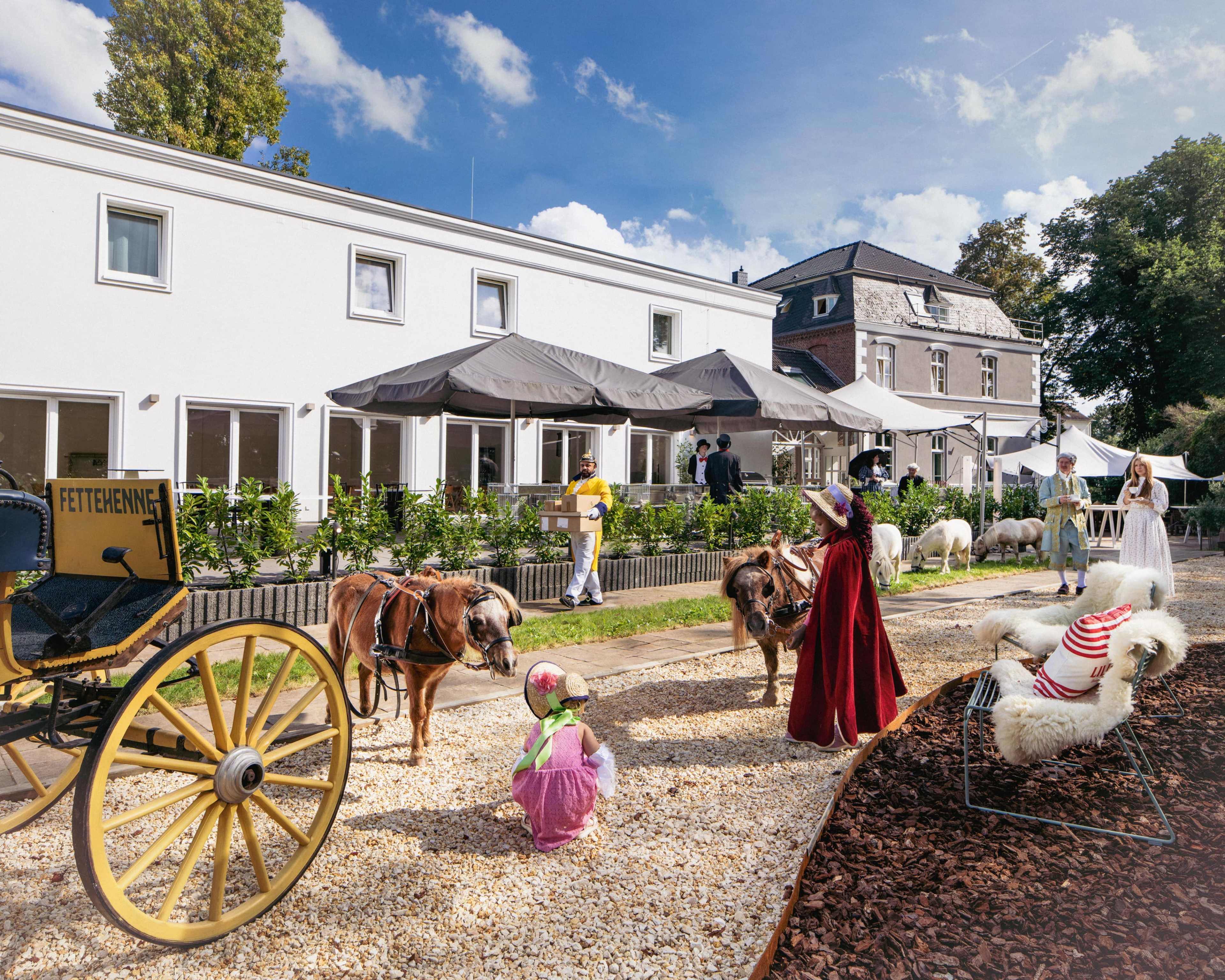 A group of people interacts with ponies and sheep in a courtyard surrounded by buildings and tables under umbrellas.