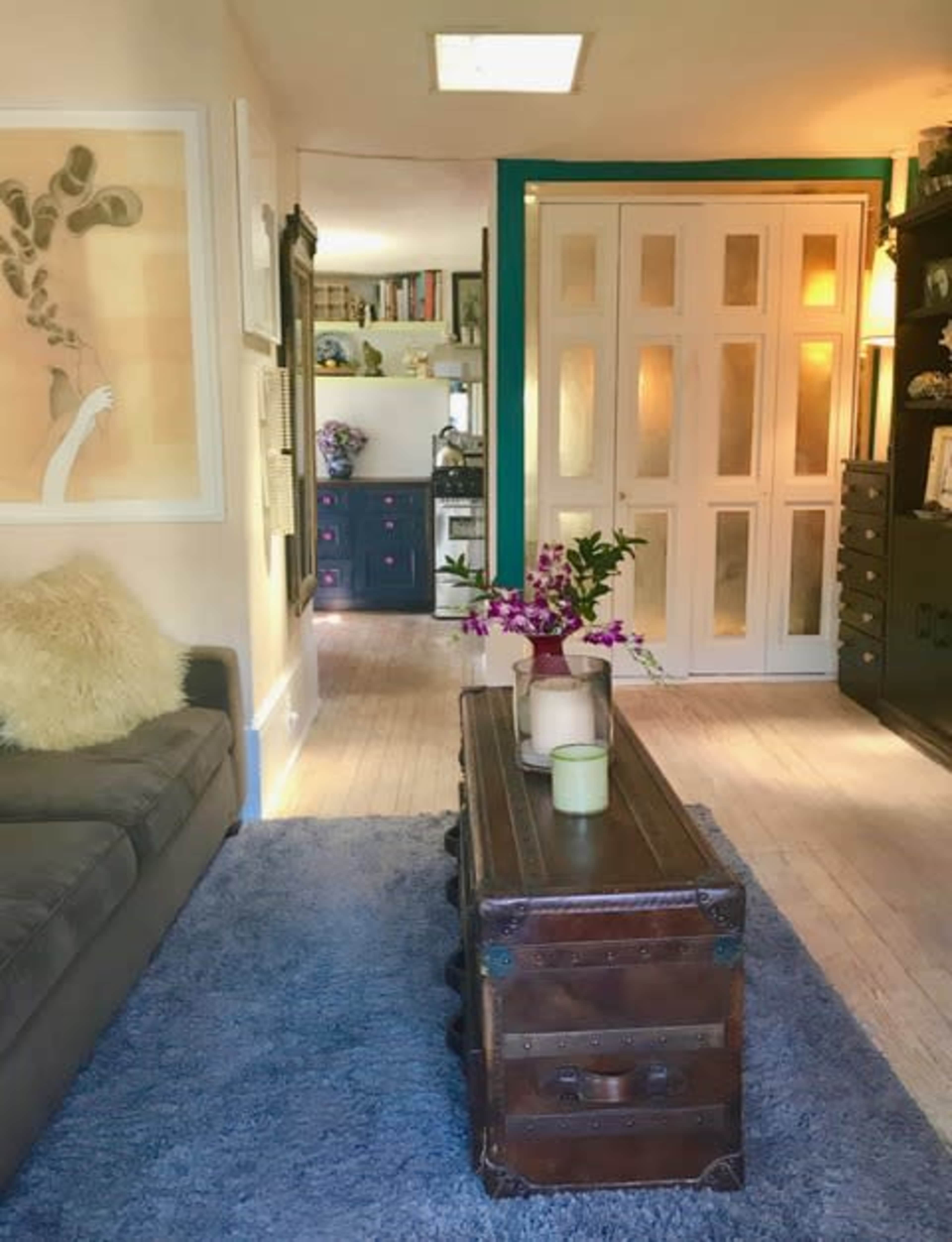 A cozy living room with a gray sofa, a wooden coffee table adorned with candles and flowers, and a view into a hallway leading to a kitchen.