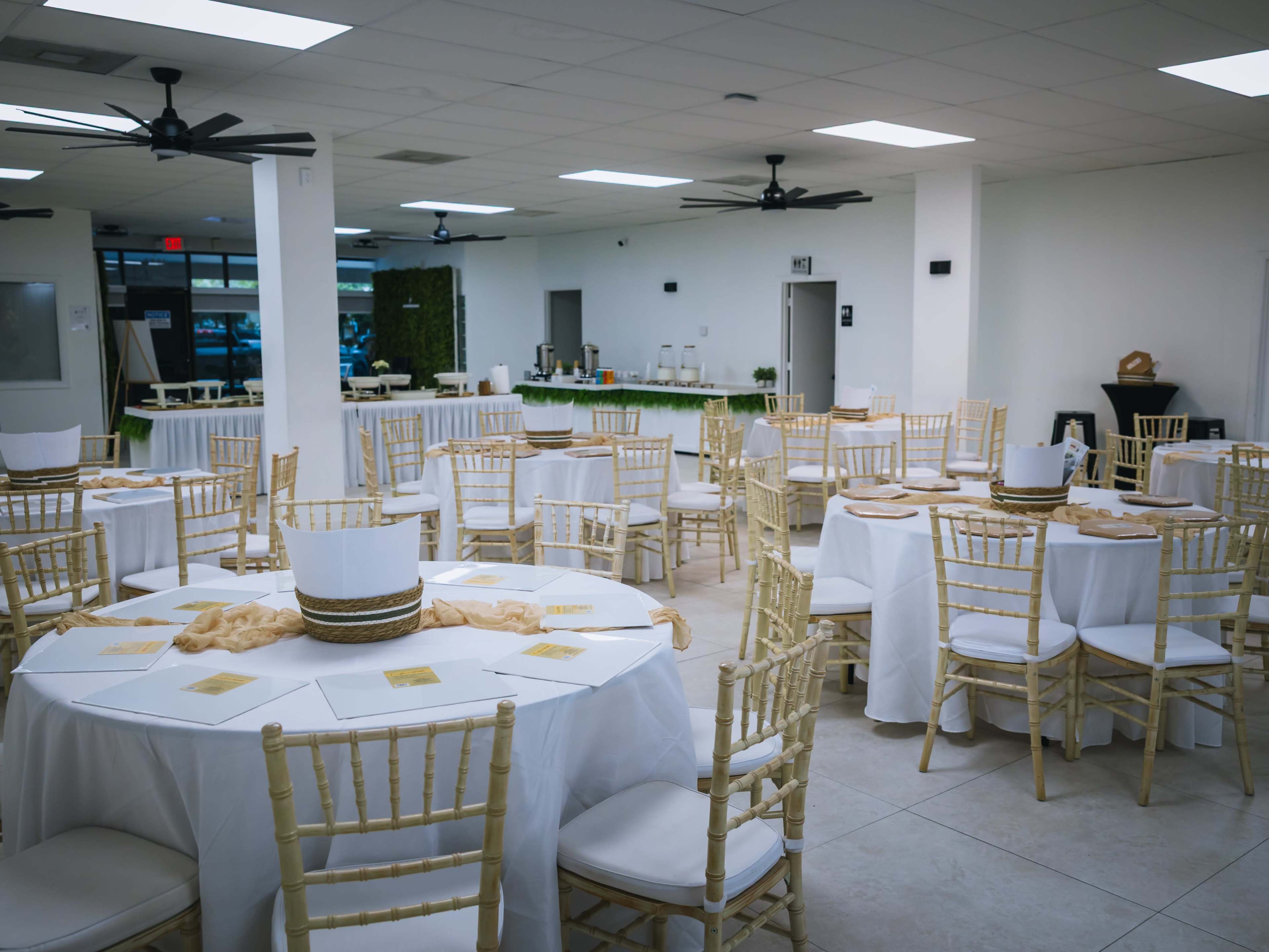 A spacious event hall features several round tables with white tablecloths, gold accents, and wooden chairs arranged in a formal setup.