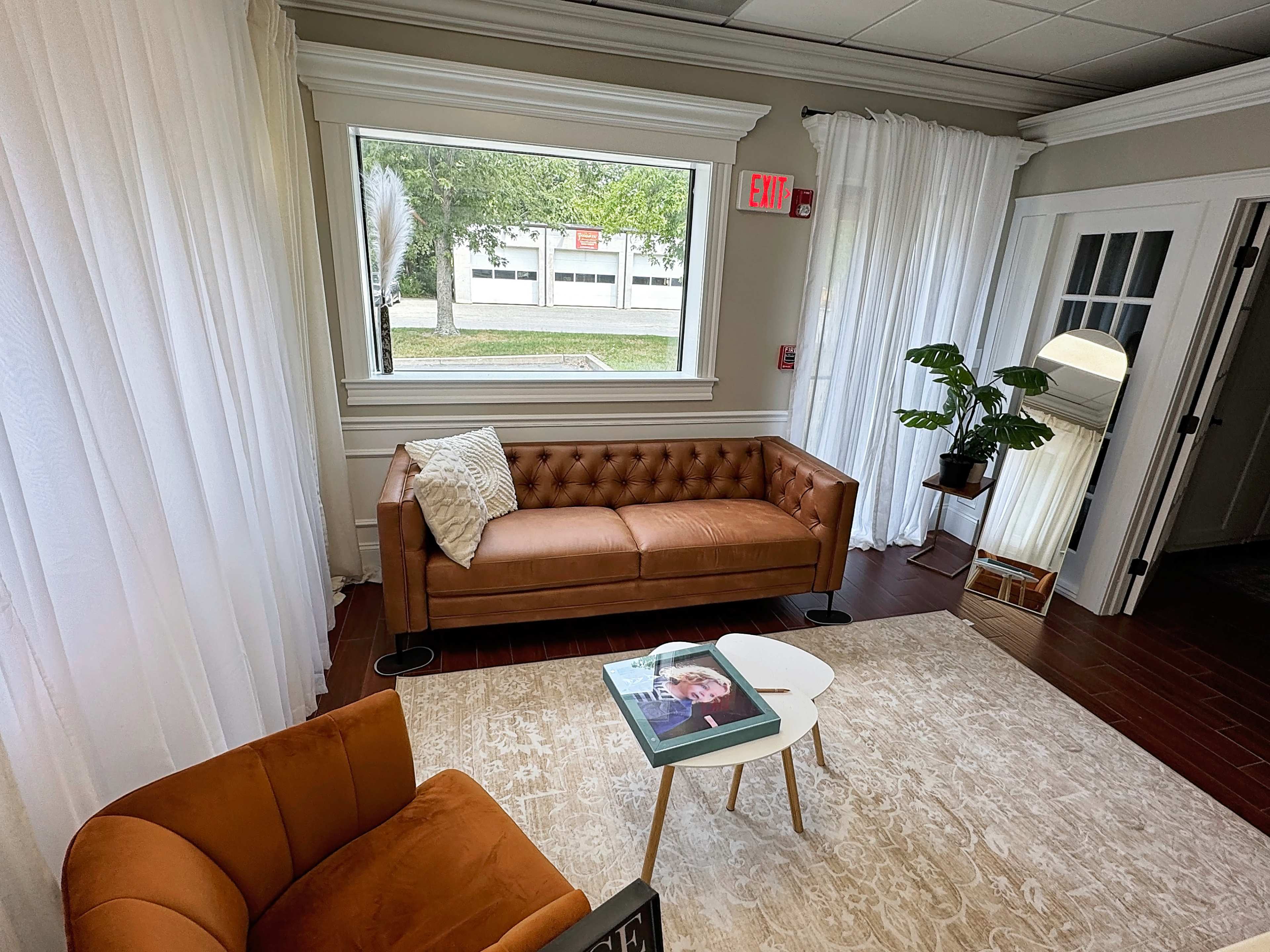 The image shows a cozy sitting area with a tan leather couch, an orange armchair, and a small coffee table, surrounded by large windows and light-colored curtains.