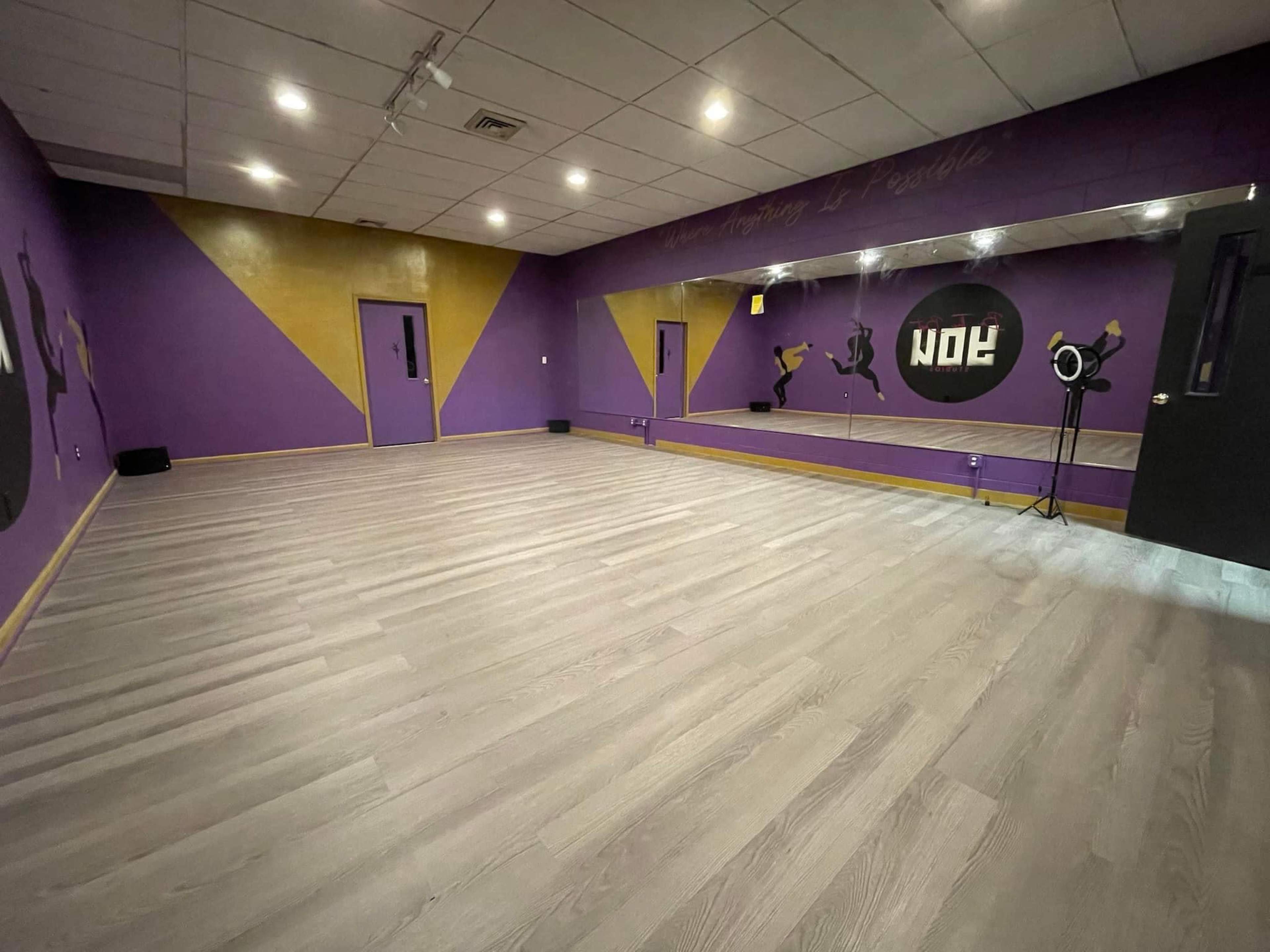 The image shows a spacious dance studio with purple and gold walls, a large mirror, and smooth wooden flooring.