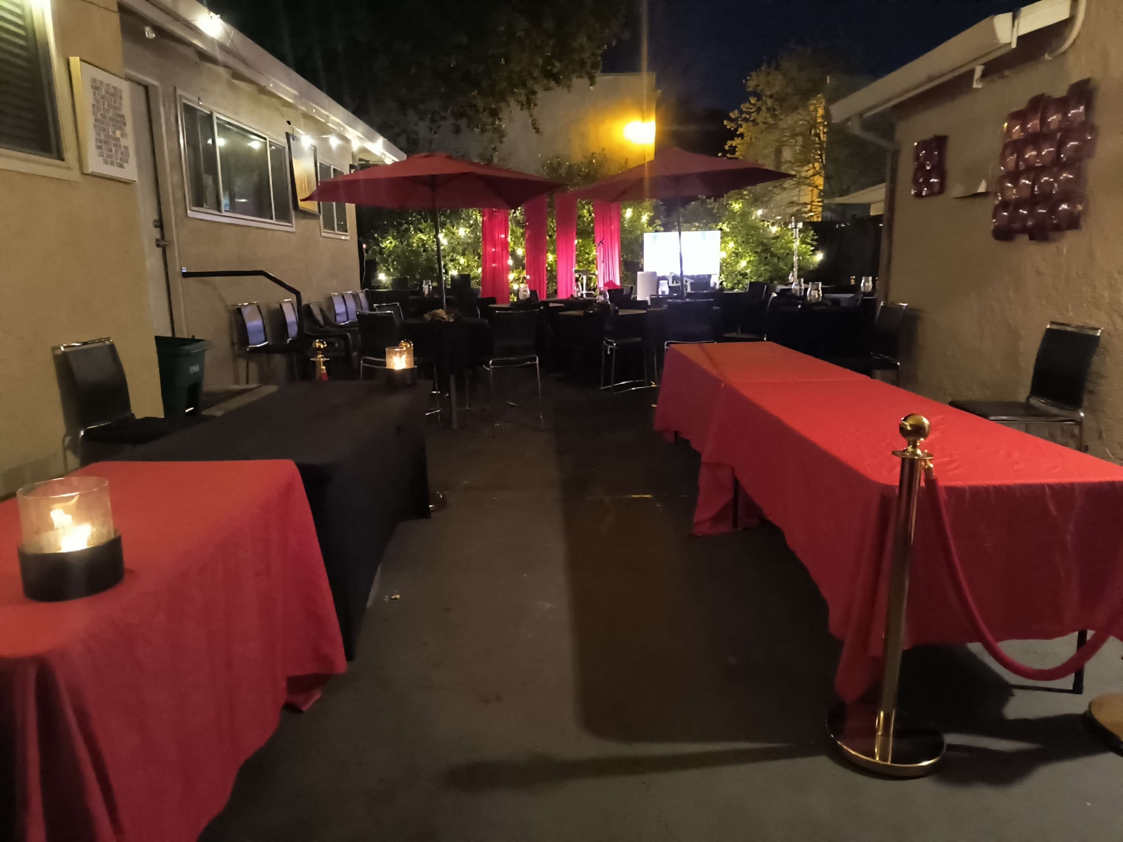 The image shows an outdoor event space with red and black tables, umbrellas, and dim lighting in the evening.