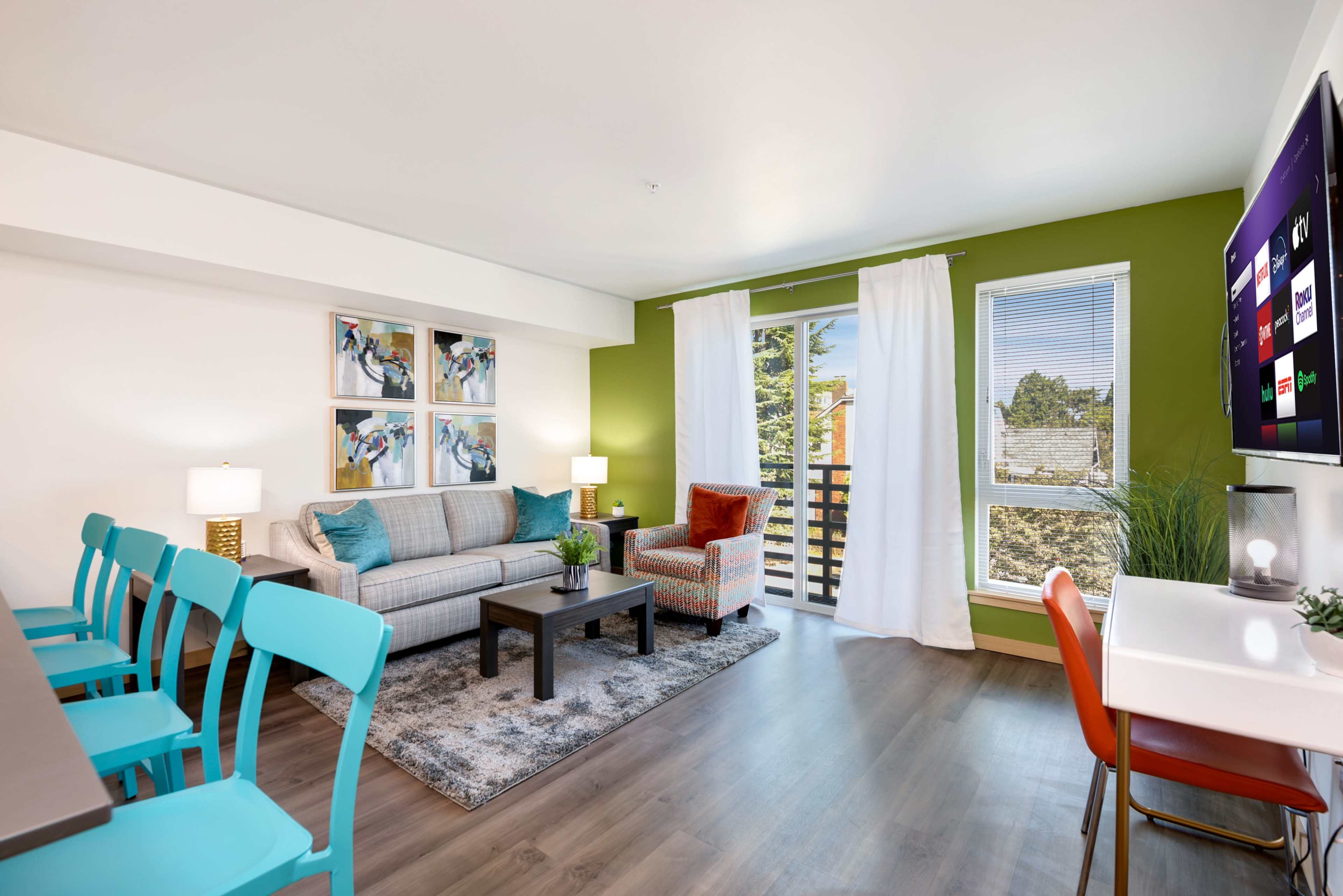 A modern living room with a sofa, coffee table, and a large TV, featuring green walls and large windows that open to a balcony.
