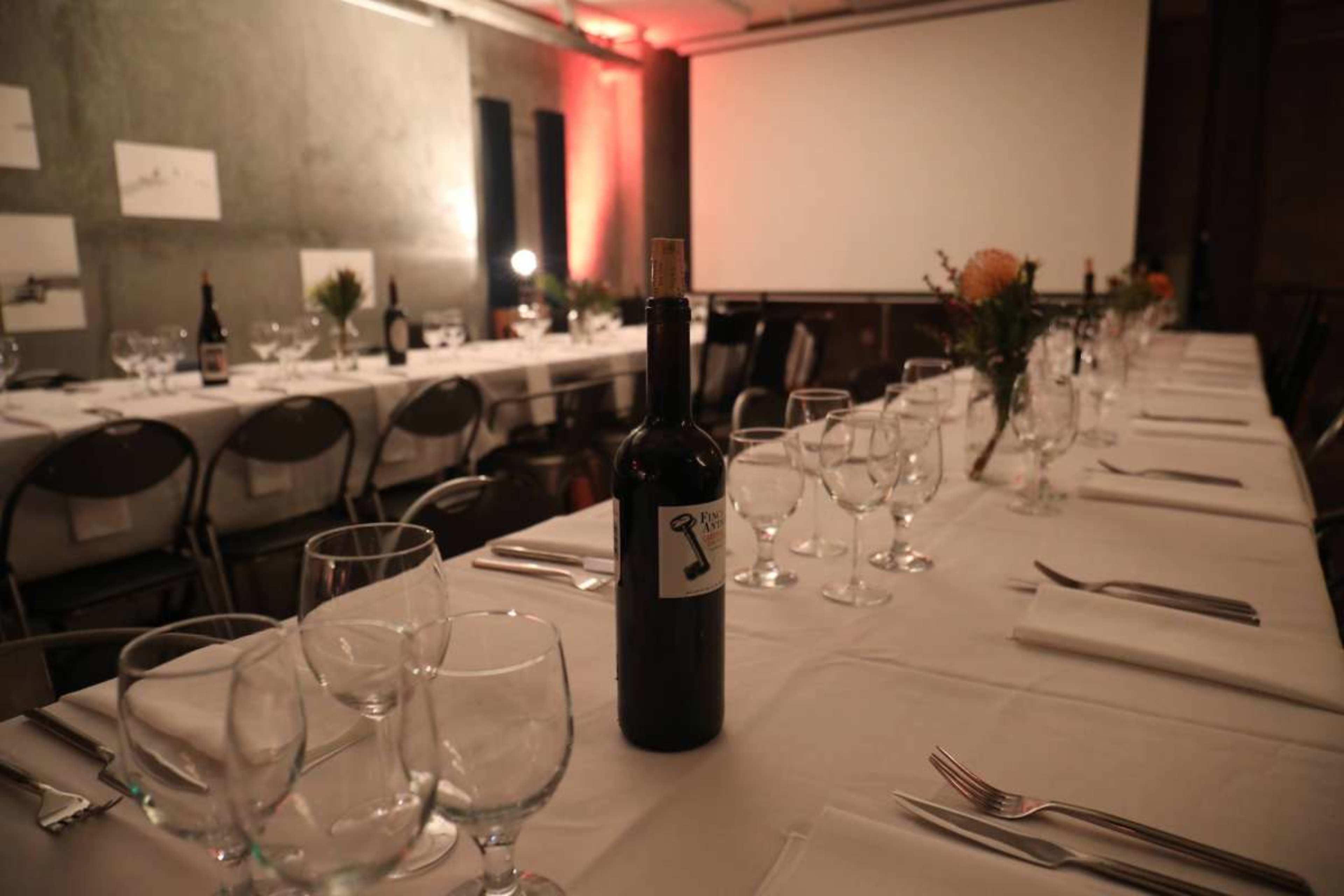 A long dining table is set with glasses and utensils, featuring a bottle of wine at the center, against a backdrop of a projection screen and dim lighting.