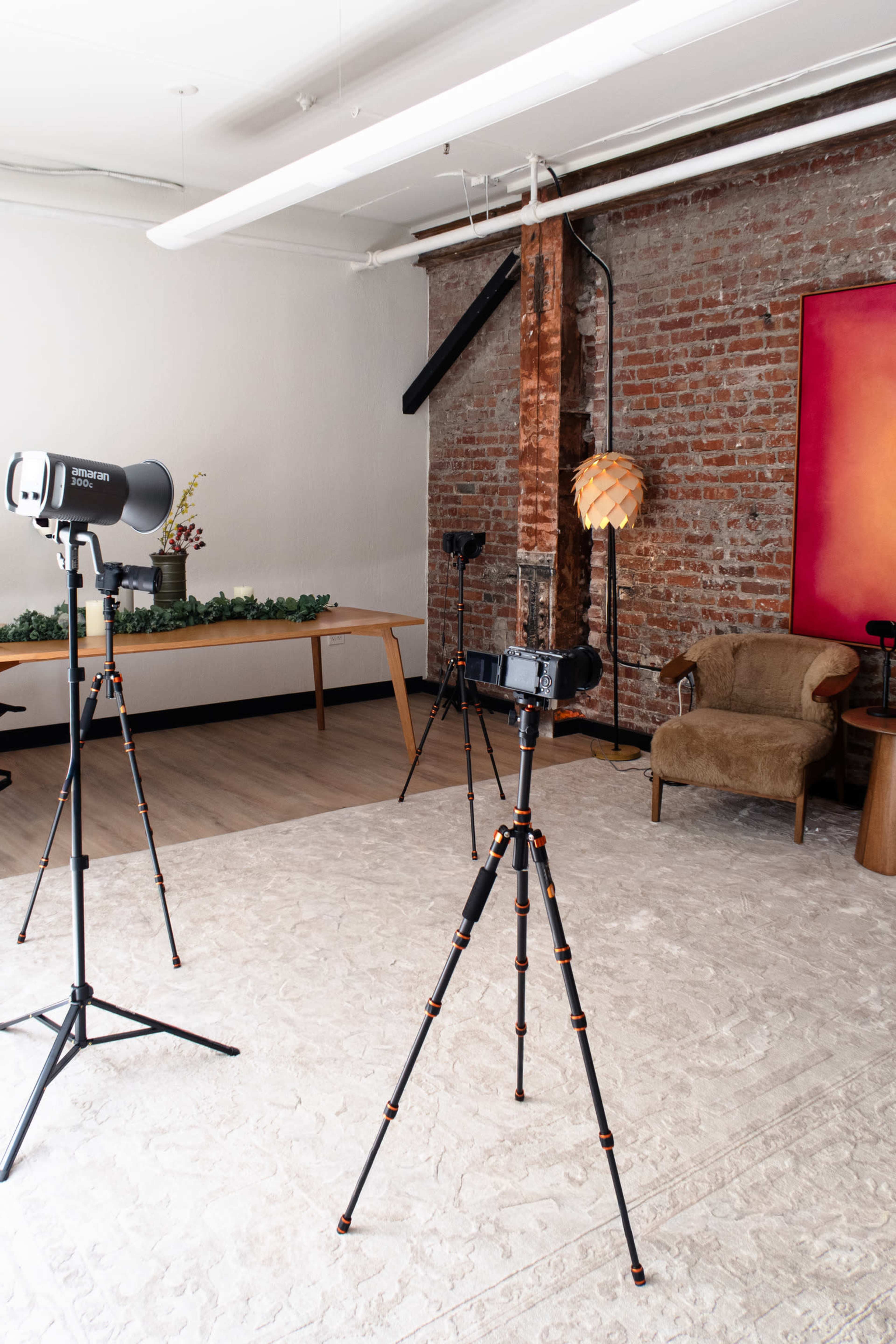 The image shows a well-lit interior space featuring two tripods with cameras, a decorative lamp, a brown chair, and a wooden table against a backdrop of exposed brick walls.