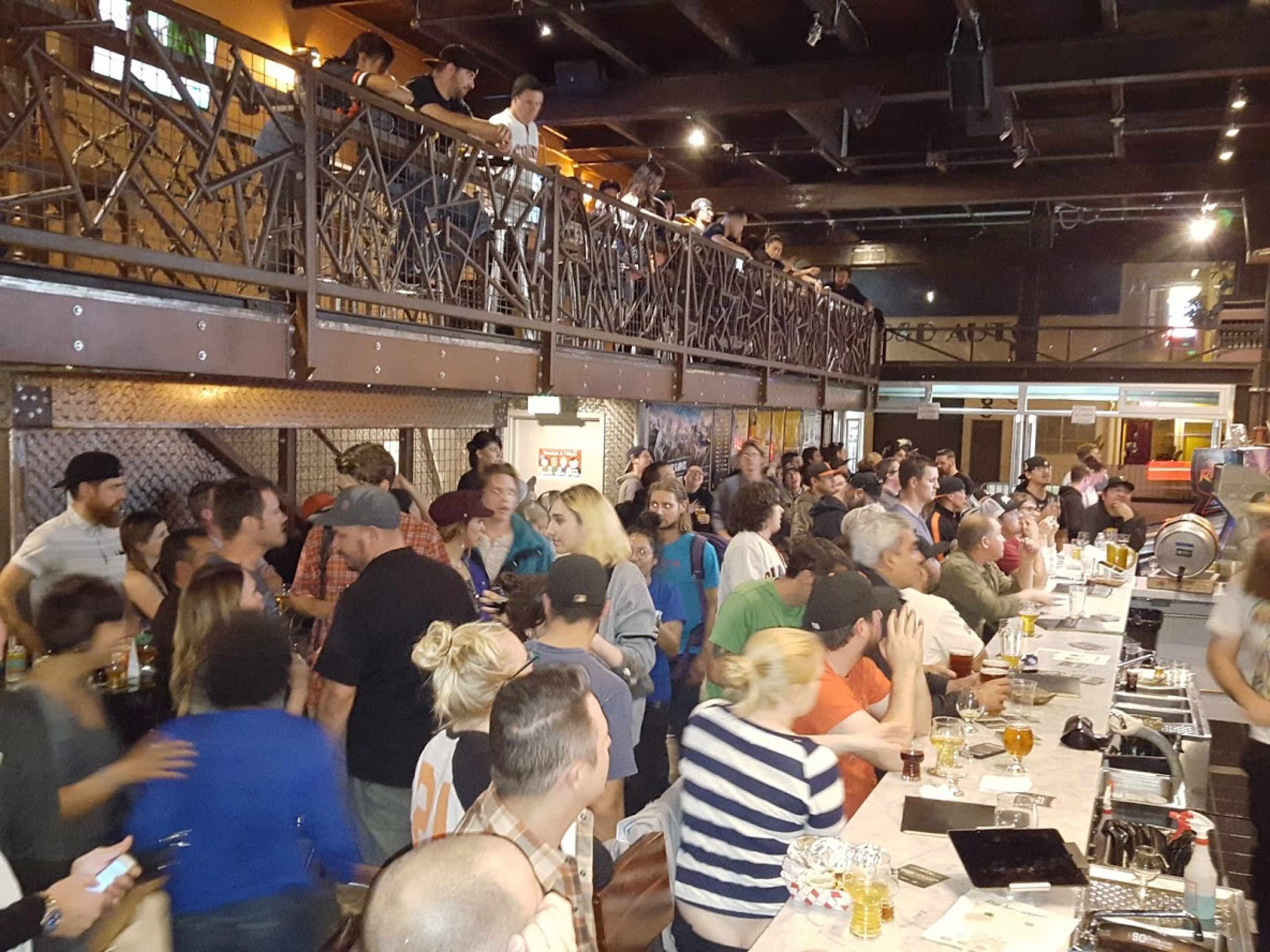 A crowded bar scene shows patrons mingling and socializing at tables and along the bar, with a second level visible in the background.