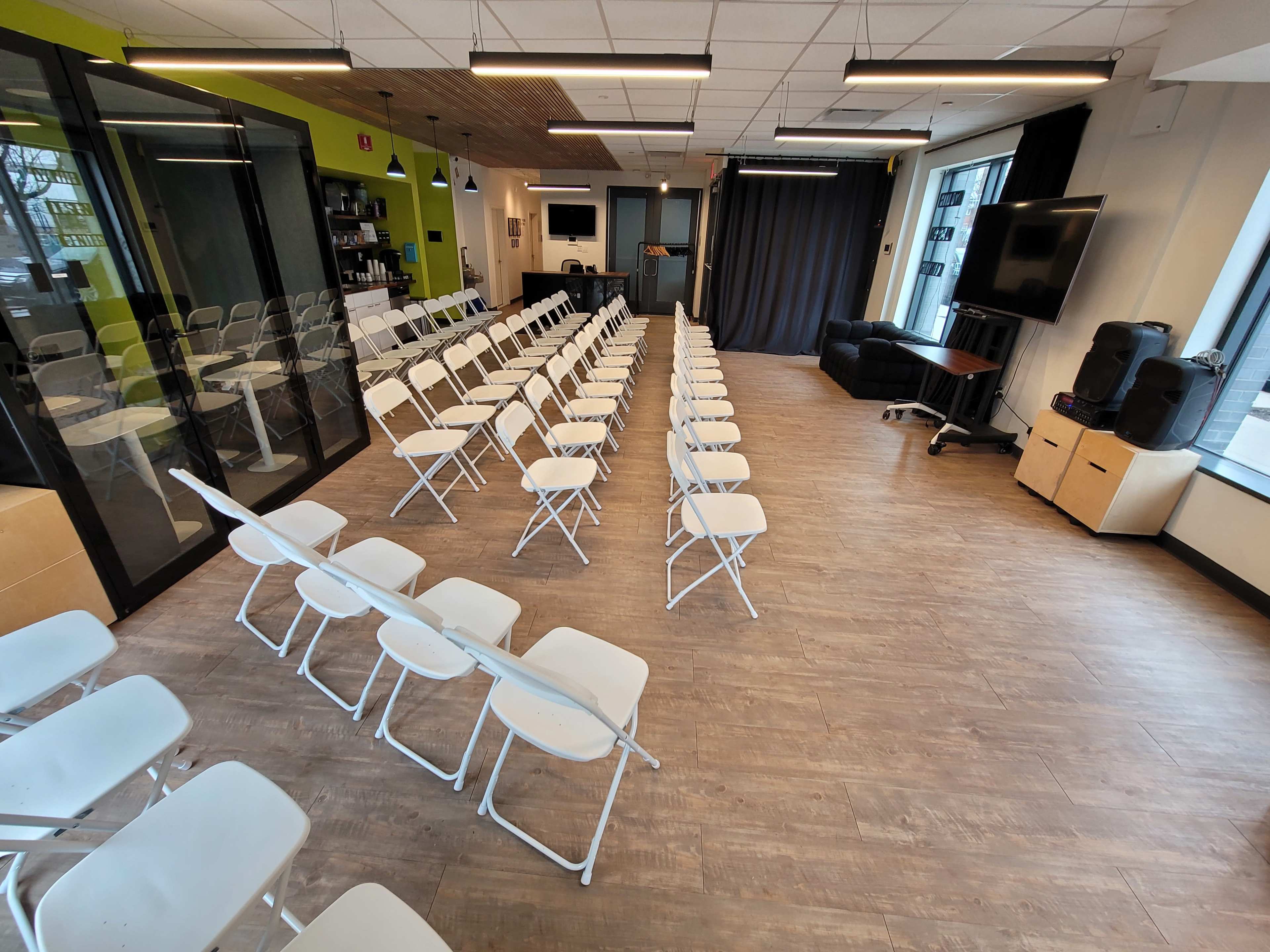 The image shows a large, open room set up with rows of white folding chairs facing a stage area, flanked by large windows and equipped with a black curtain and a television.