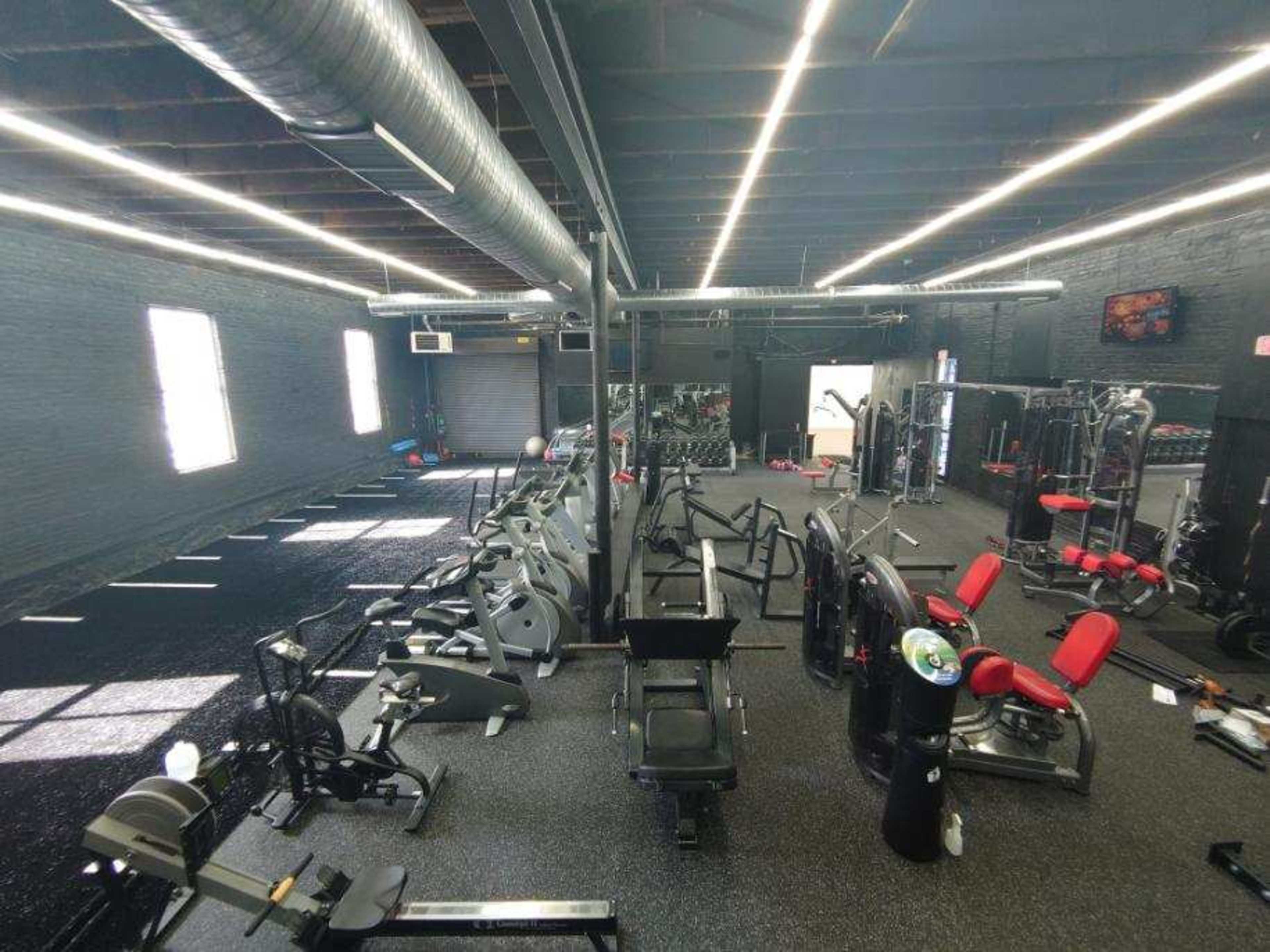 The image shows a fitness gym interior with various exercise machines and equipment arranged neatly along the space, illuminated by overhead lighting.