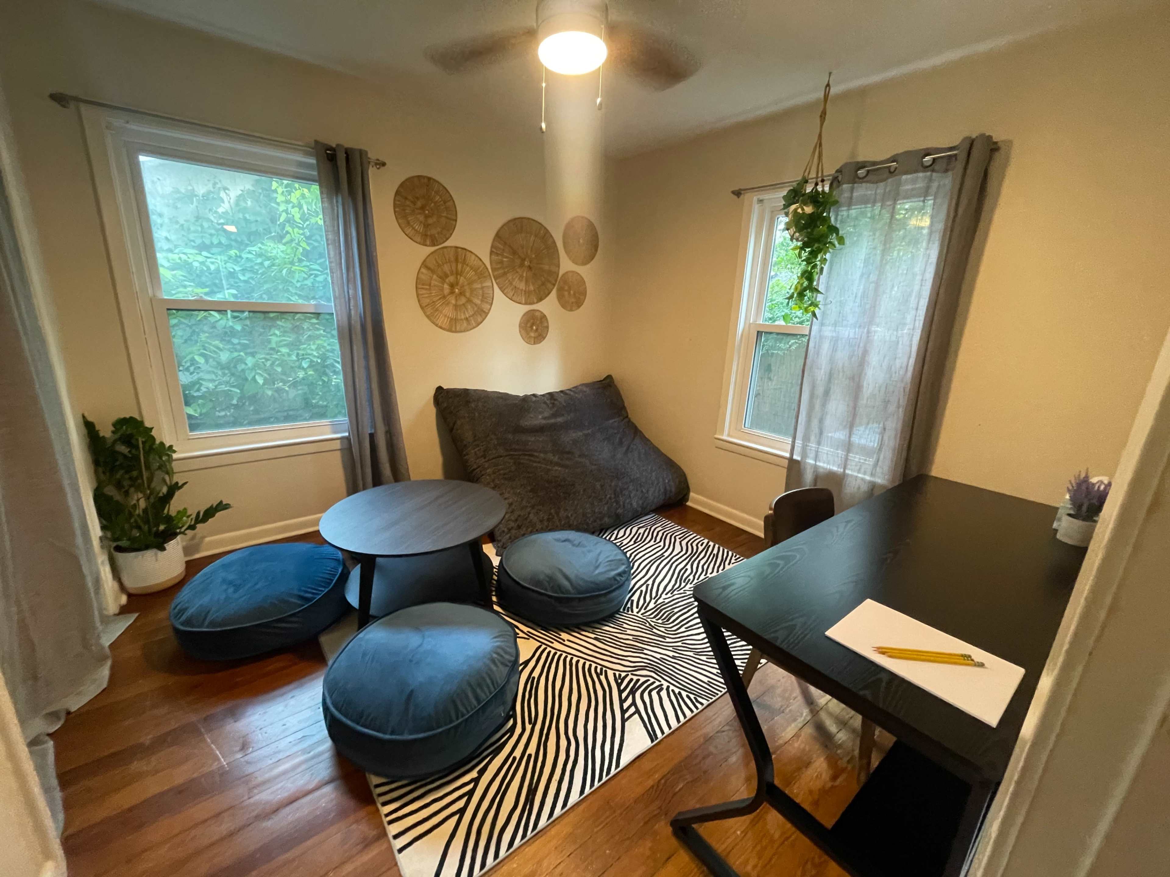 The image shows a cozy room with a large bean bag chair, two round ottomans, a small table, a desk, and decorative wall pieces near the windows.