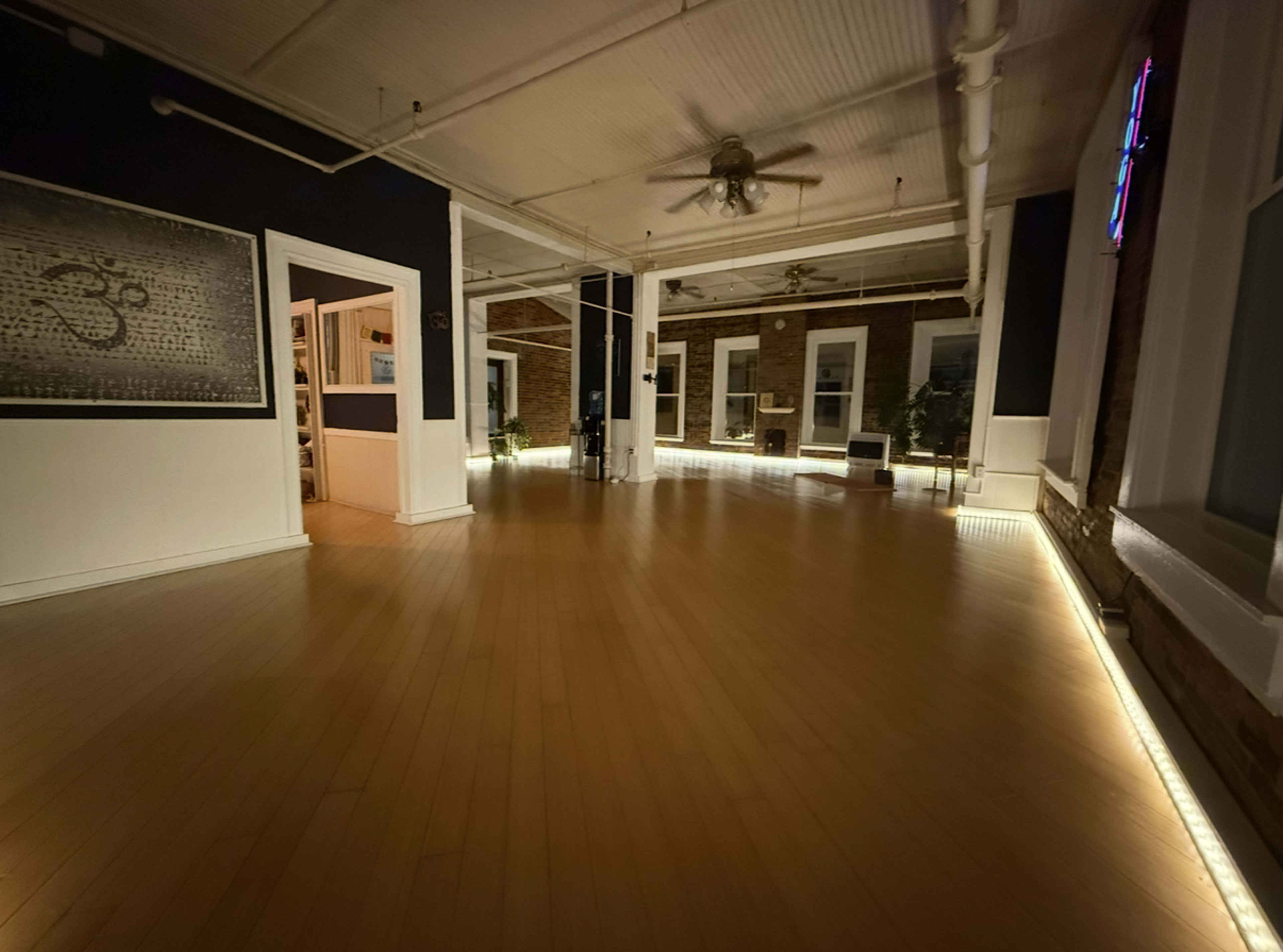 The image shows a spacious indoor room with wooden flooring, illuminated by soft lighting along the edges and featuring brick walls and decorative plants.