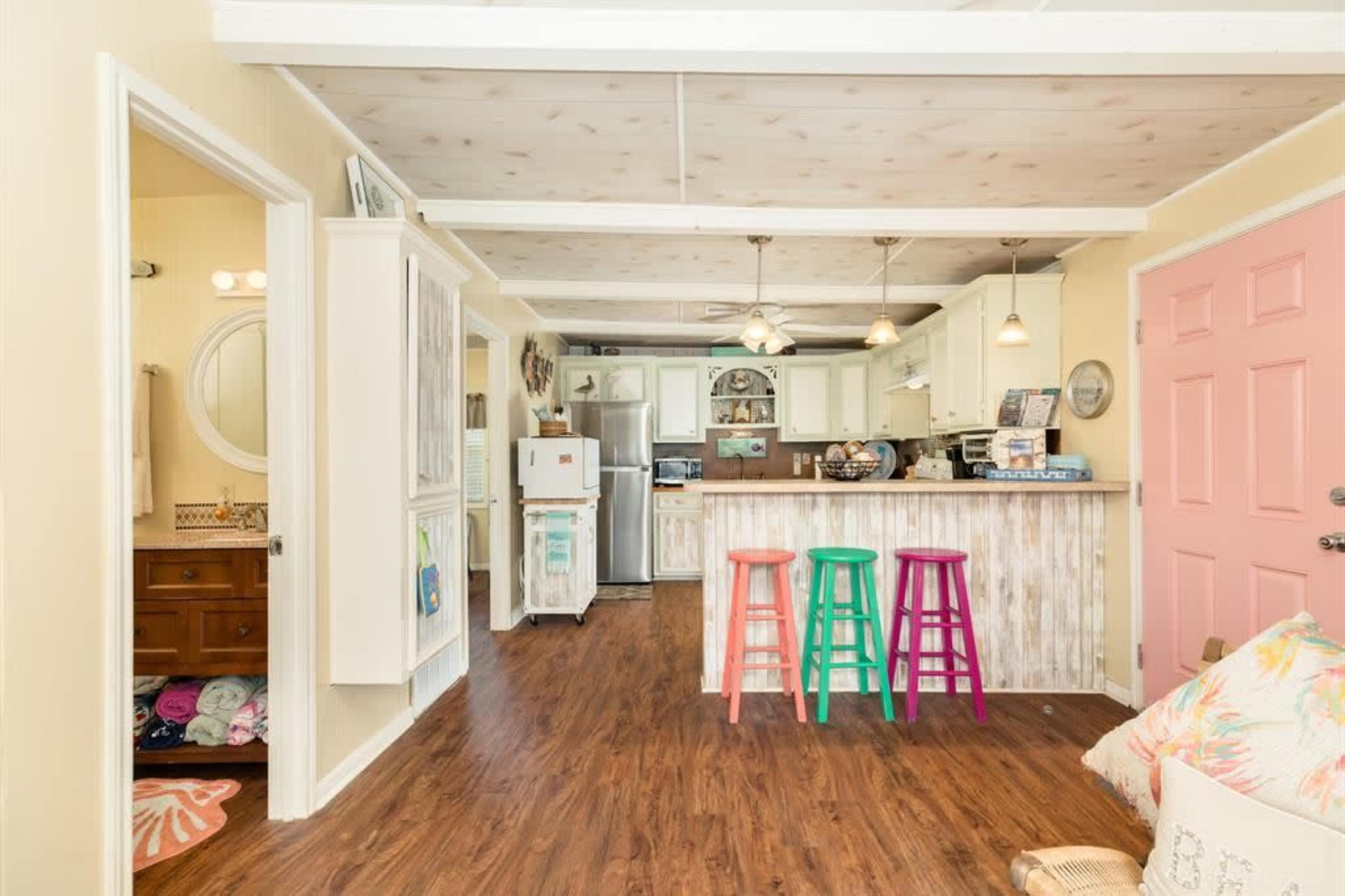 The image shows a bright, open kitchen and living area featuring a bar with three colorful stools, a pink door, and light-colored cabinets.