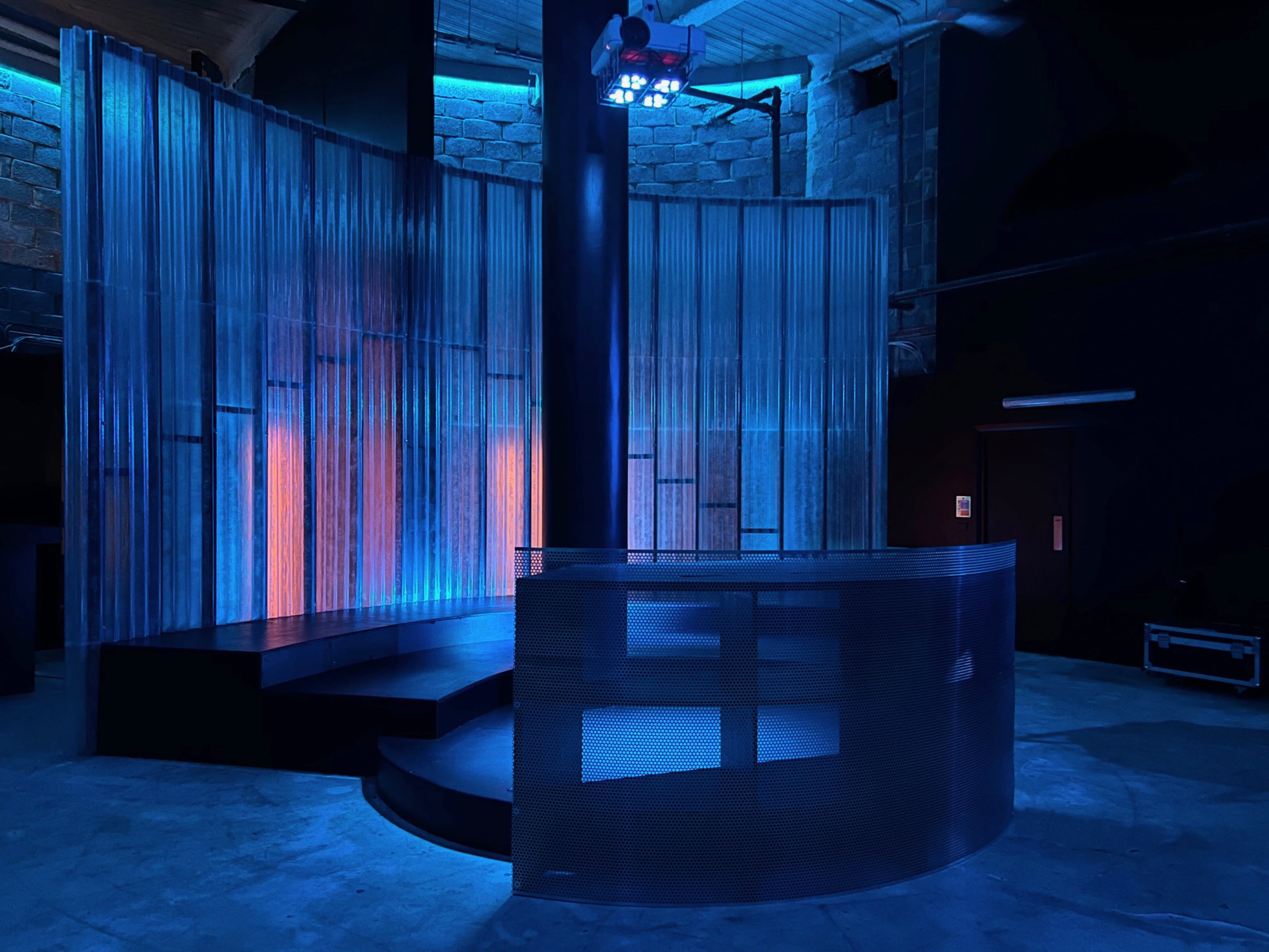 The image shows a dimly lit modern stage with a curved metal bar and textured walls, illuminated by blue and orange lights.