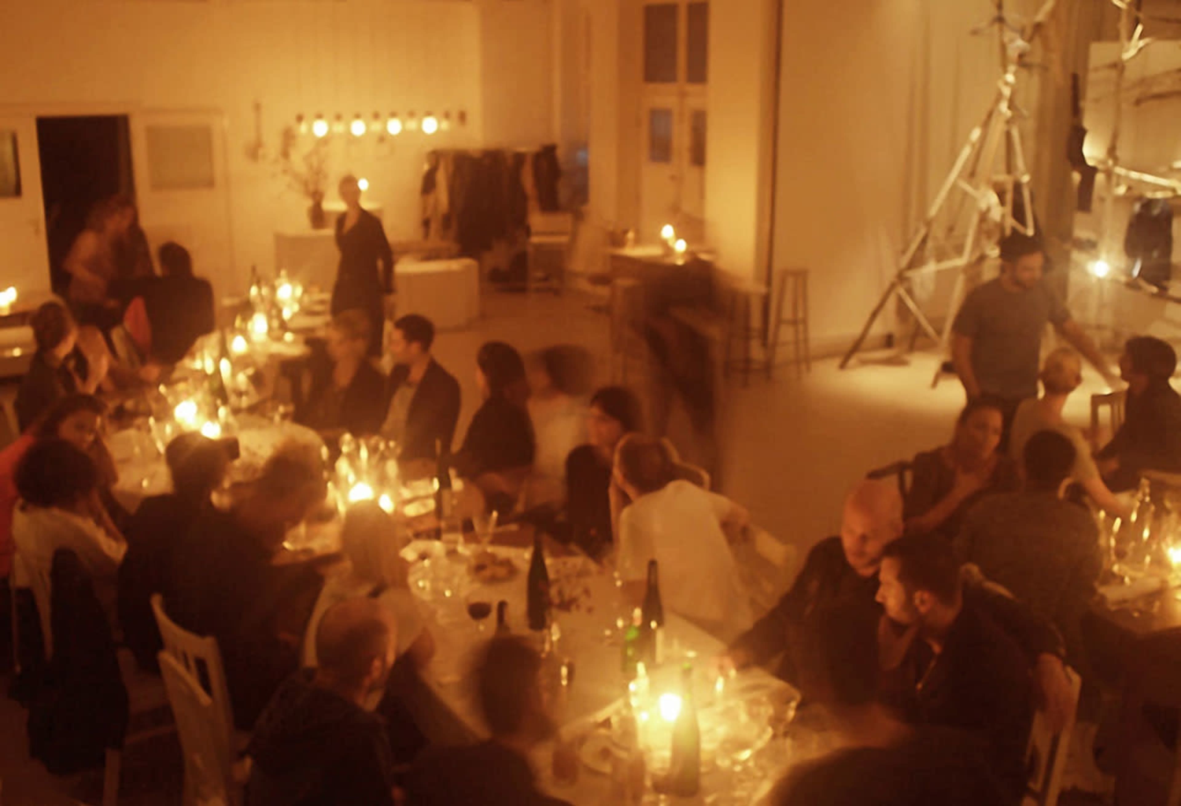 A dimly lit dining space is filled with people gathered around long tables adorned with candles and plates during a festive gathering.