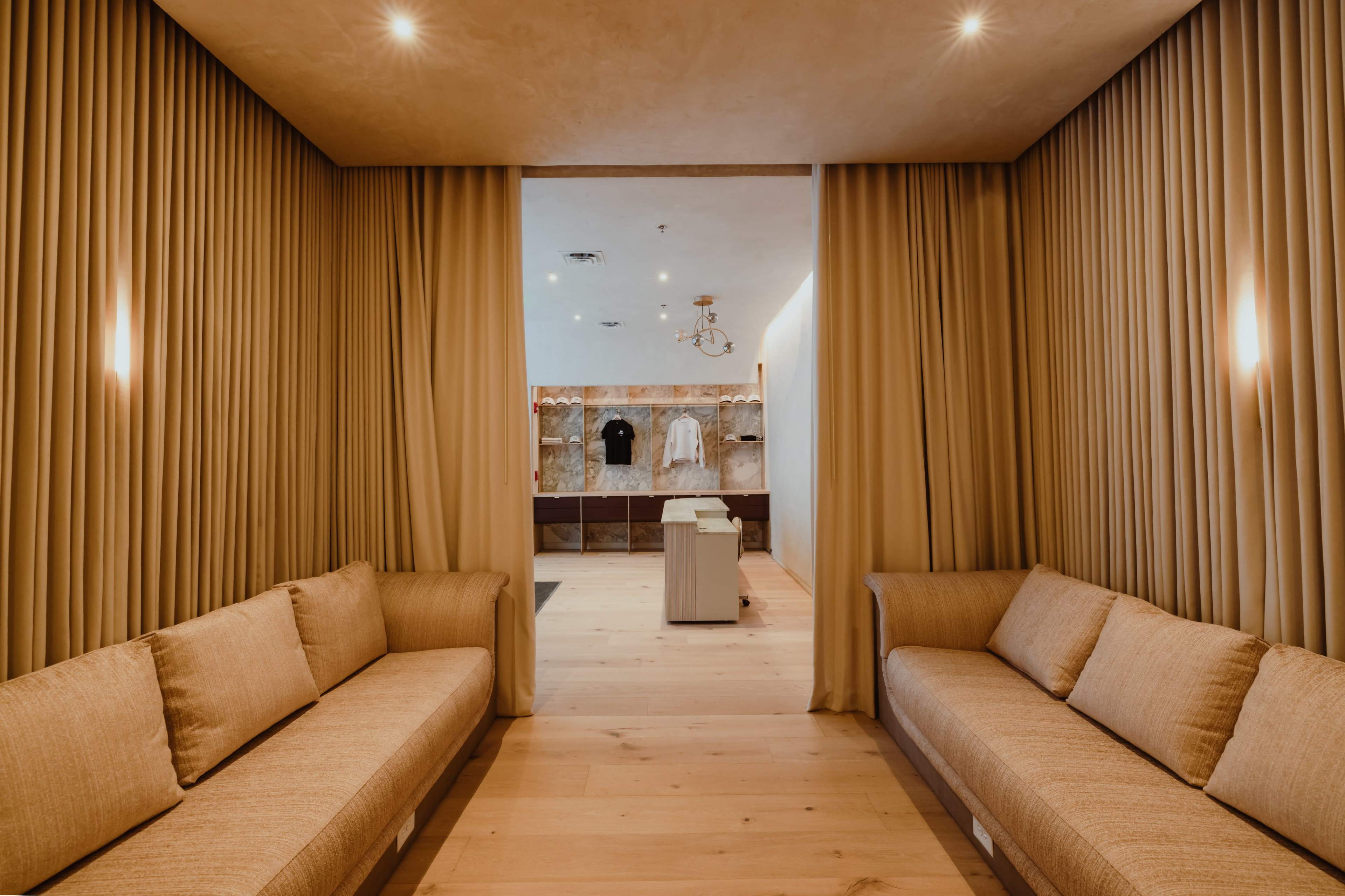 The image shows a warm-toned interior with two beige couches positioned opposite each other, framed by flowing curtains, leading into a brighter space with clothing displays.