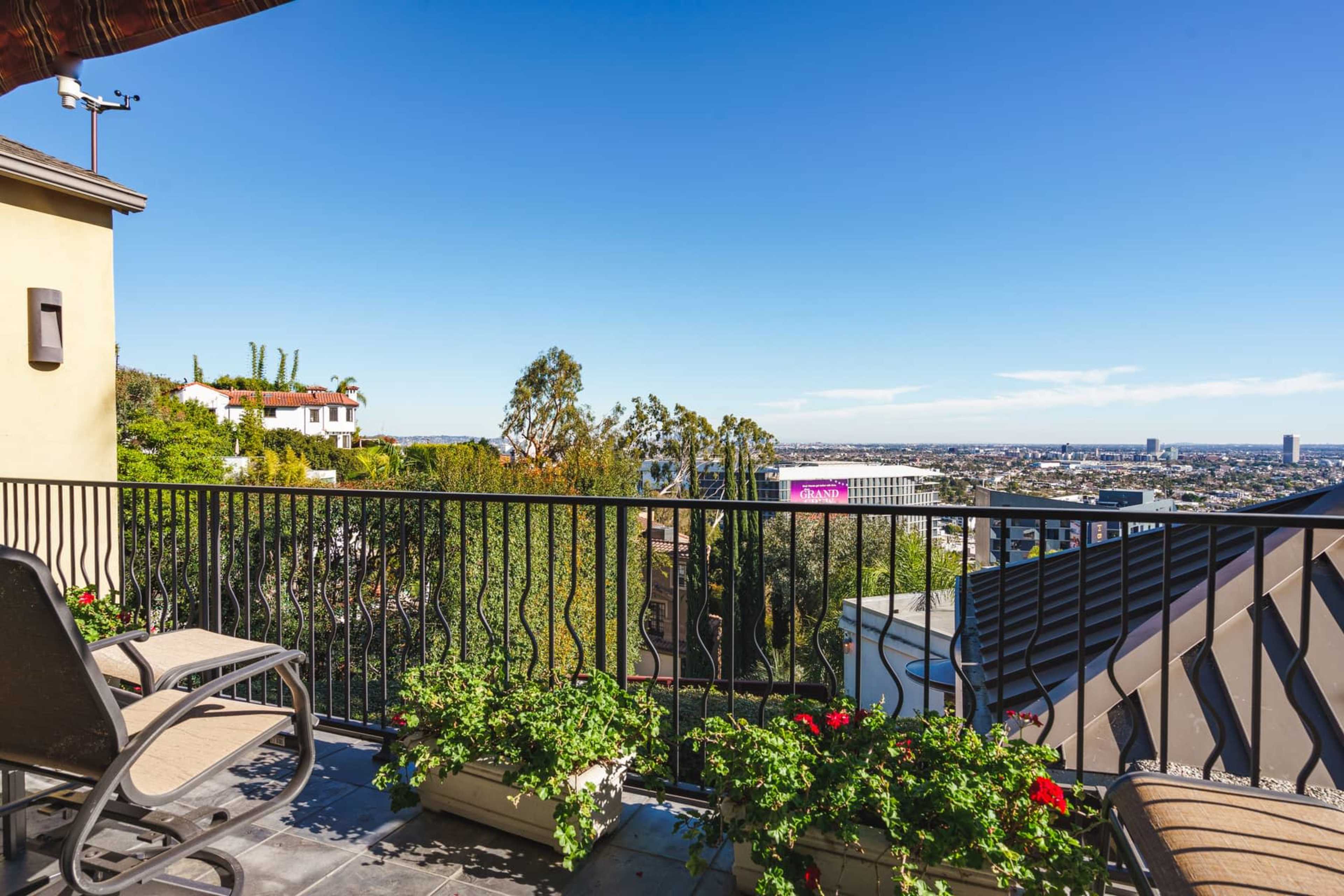The image shows a balcony with outdoor seating and potted flowers overlooking a city landscape under a clear sky.