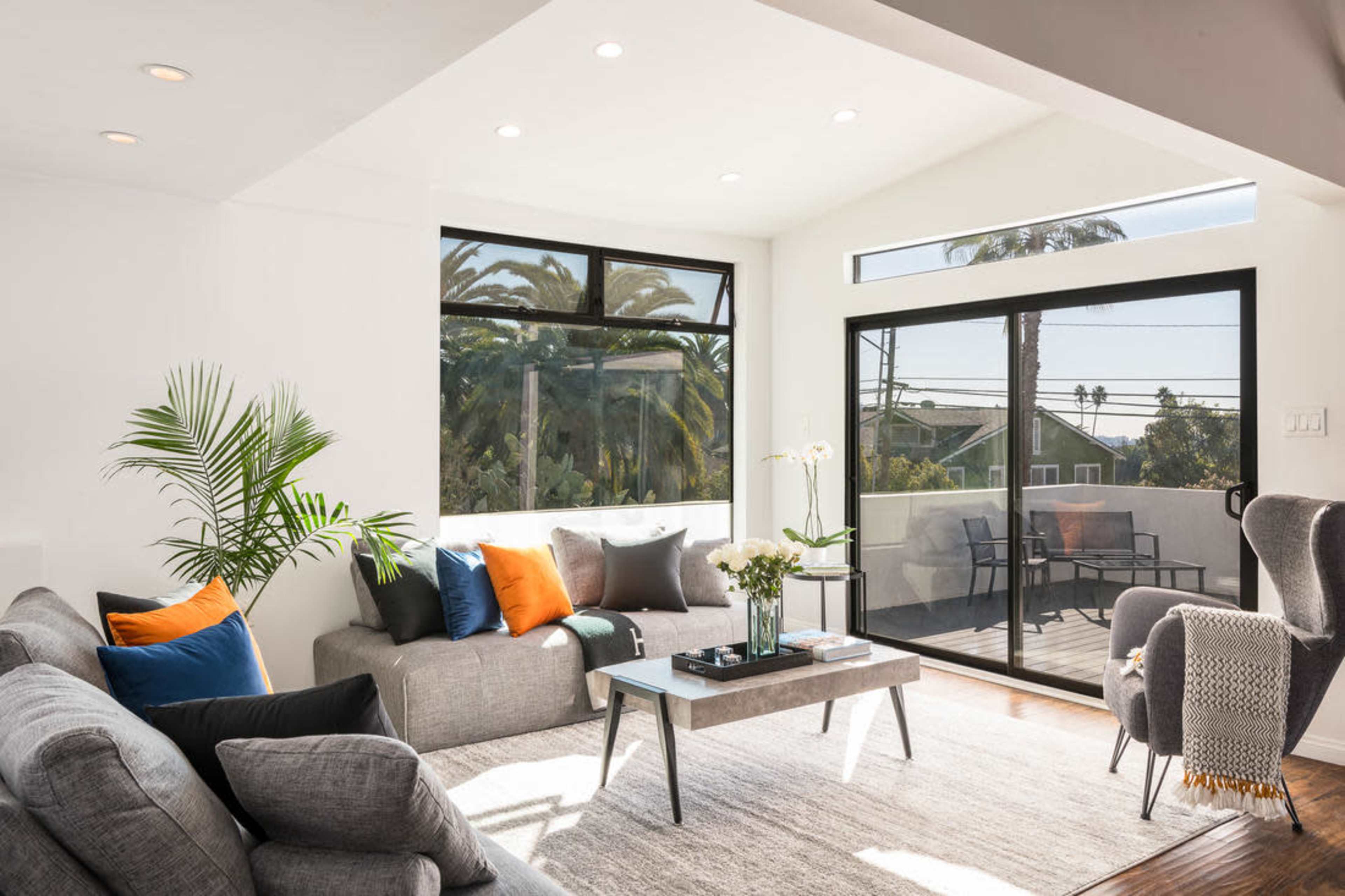 A modern living room with large windows showcasing a view of palm trees, furnished with gray sofas and a coffee table.